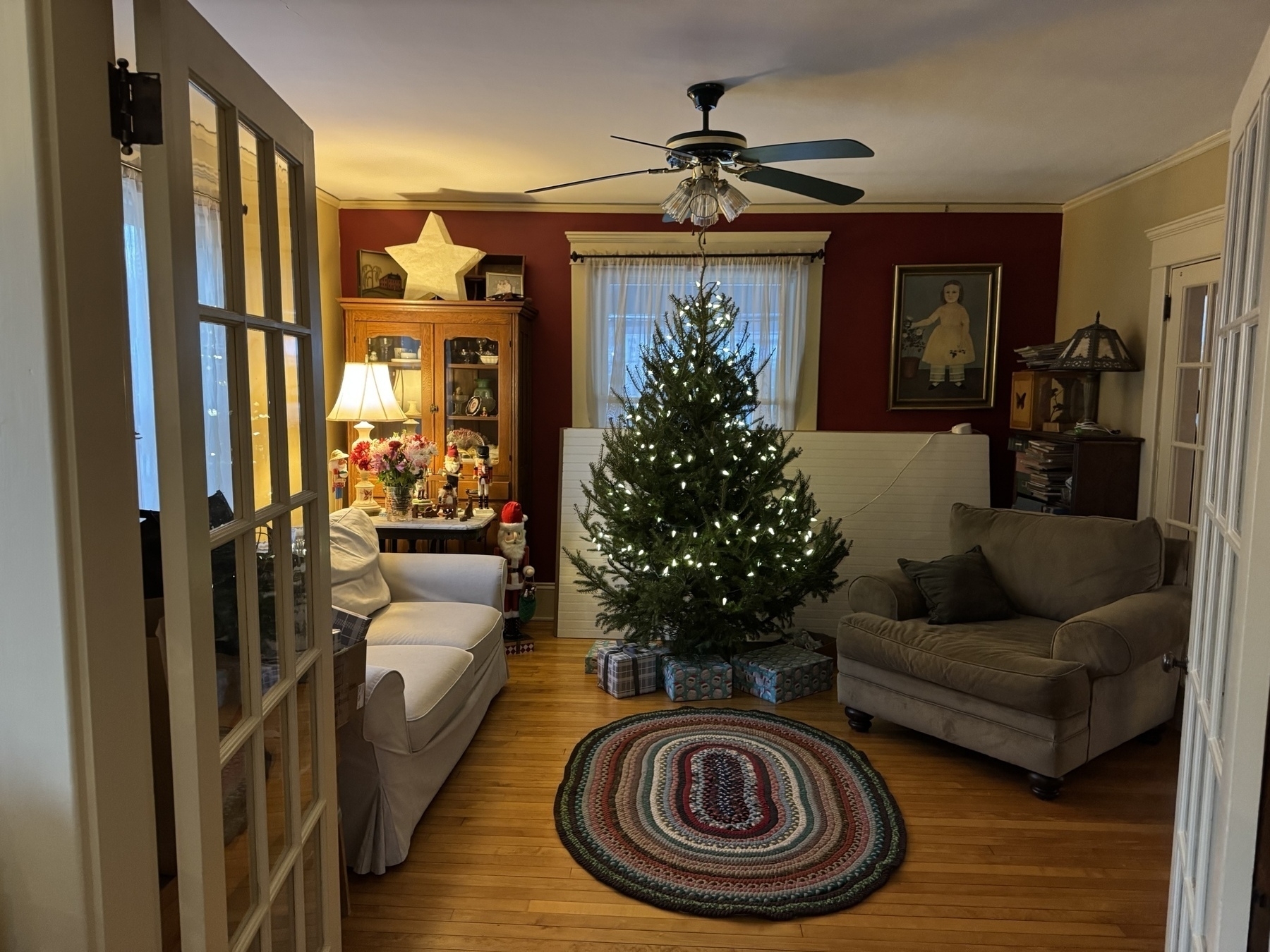 A cozy living room is decorated for the holidays with a lit Christmas tree, presents underneath, and warm ambient lighting.