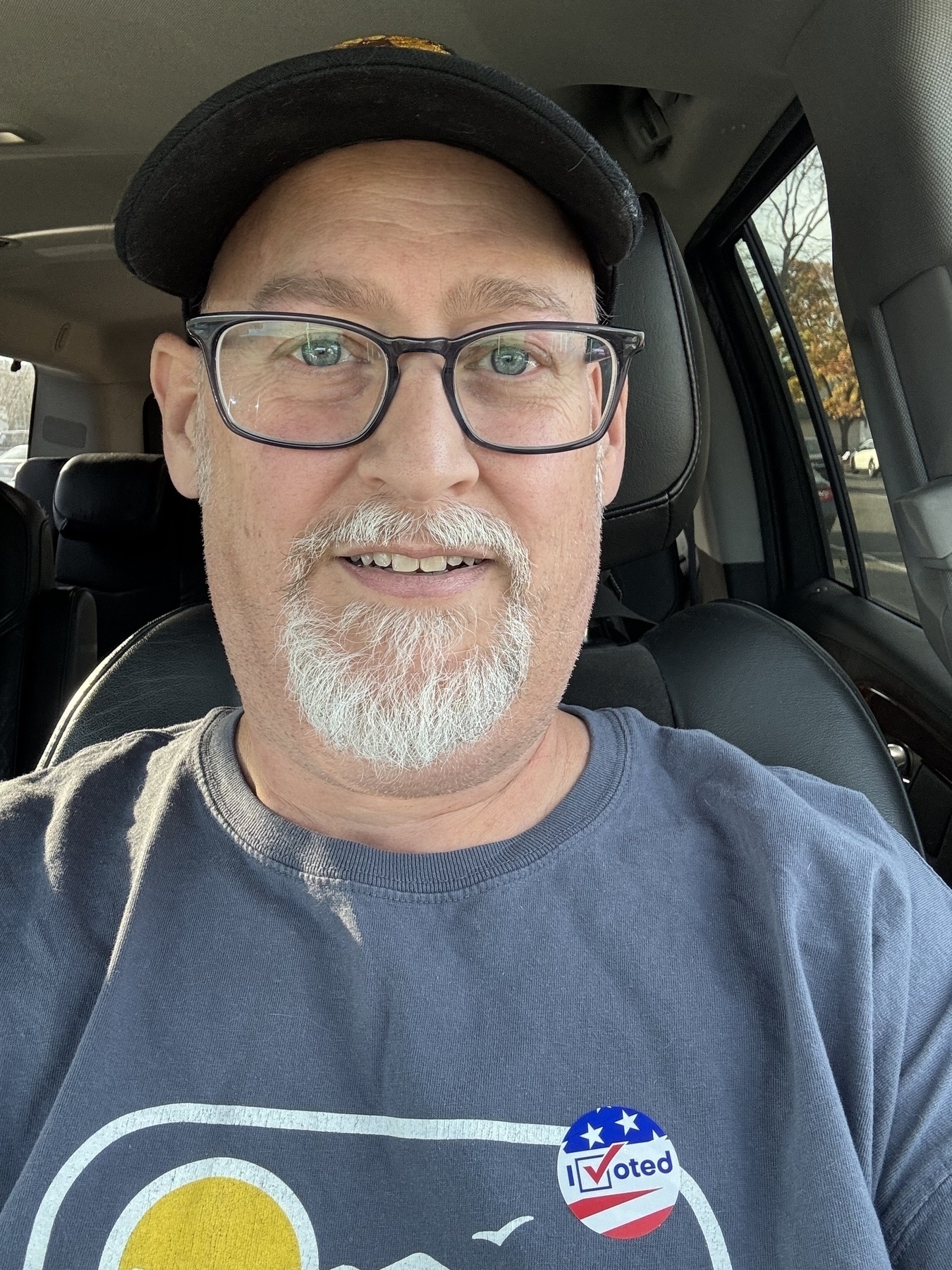 Me with glasses and a goatee, wearing a cap and a t-shirt with an "I Voted" sticker, is sitting in a car.