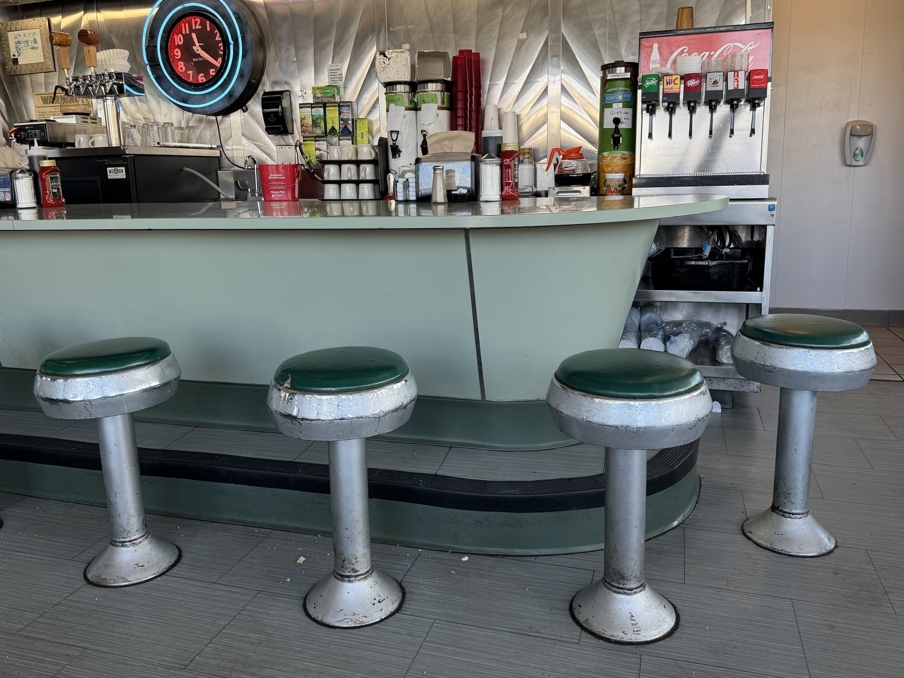 A retro-style diner counter features four green stools, a soda fountain, and a vintage clock on the wall.