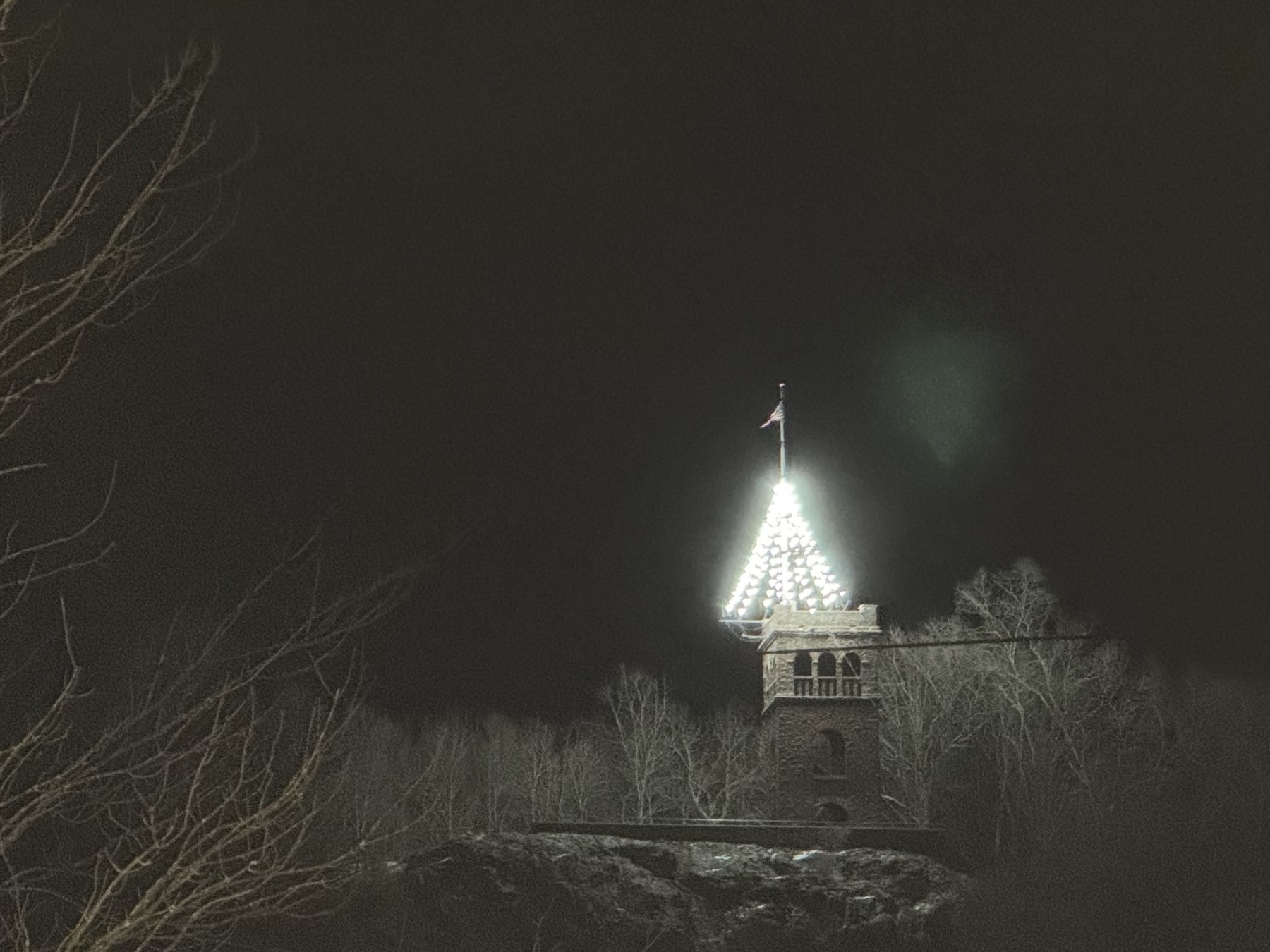 A stone tower with a brightly illuminated triangular lights stands amidst bare trees at night.