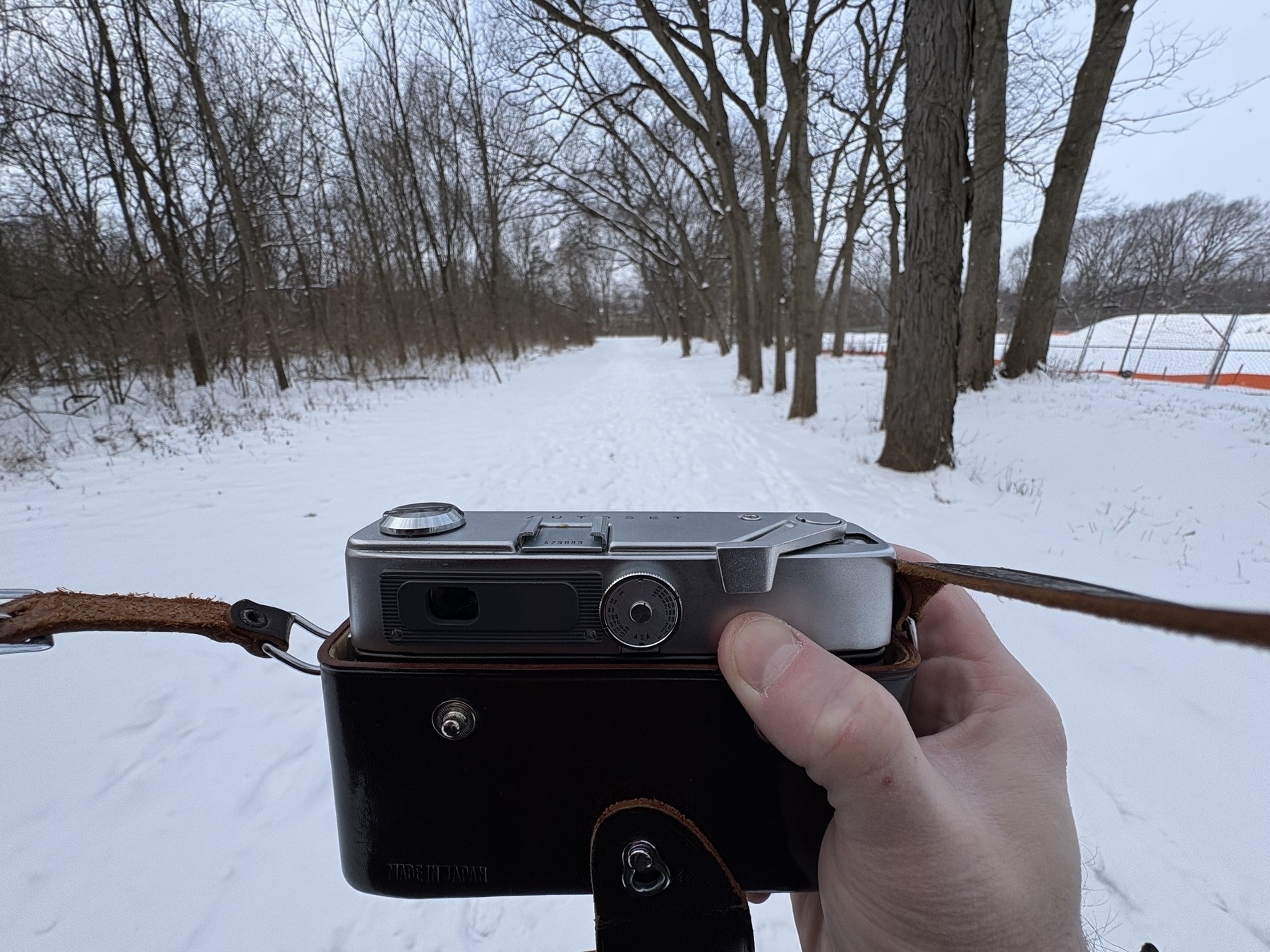 Auto-generated description: A hand holding a vintage camera is capturing a snowy, tree-lined path.