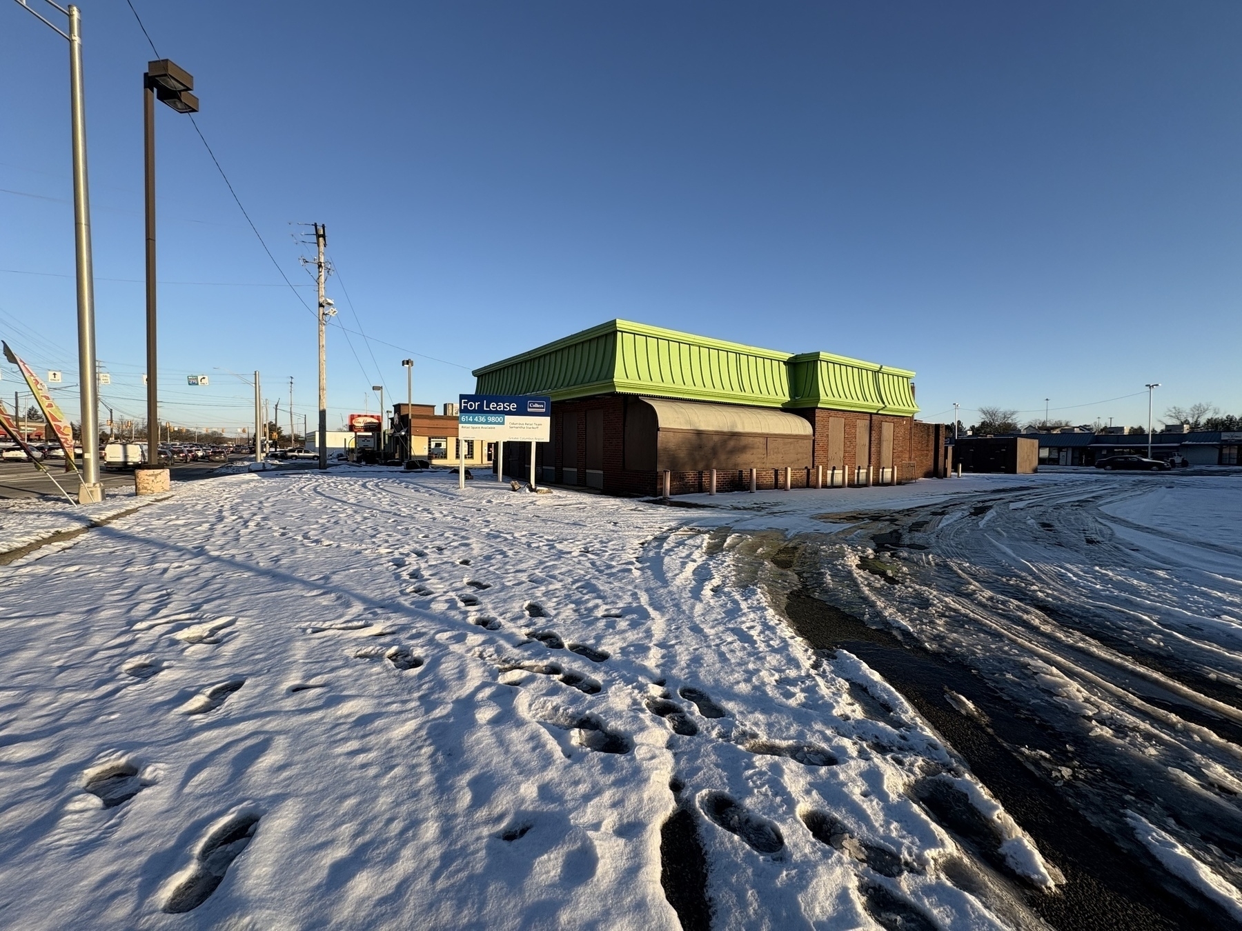 Auto-generated description: A snow-covered lot with footprints leads to an empty, green-roofed building available for lease, under a clear blue sky.