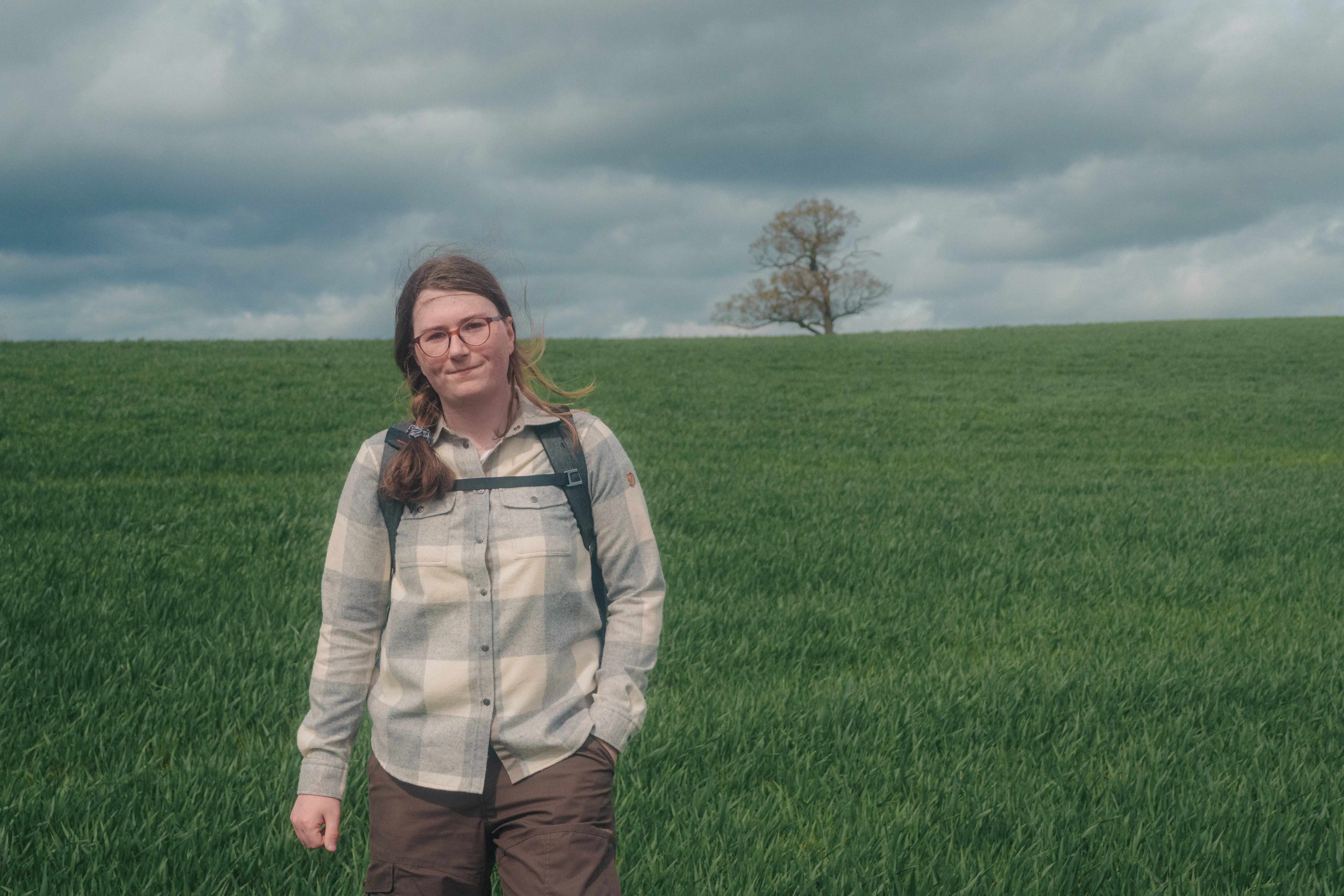 A person wearing a backpack stands in a grassy field with a tree and a cloudy sky in the background.