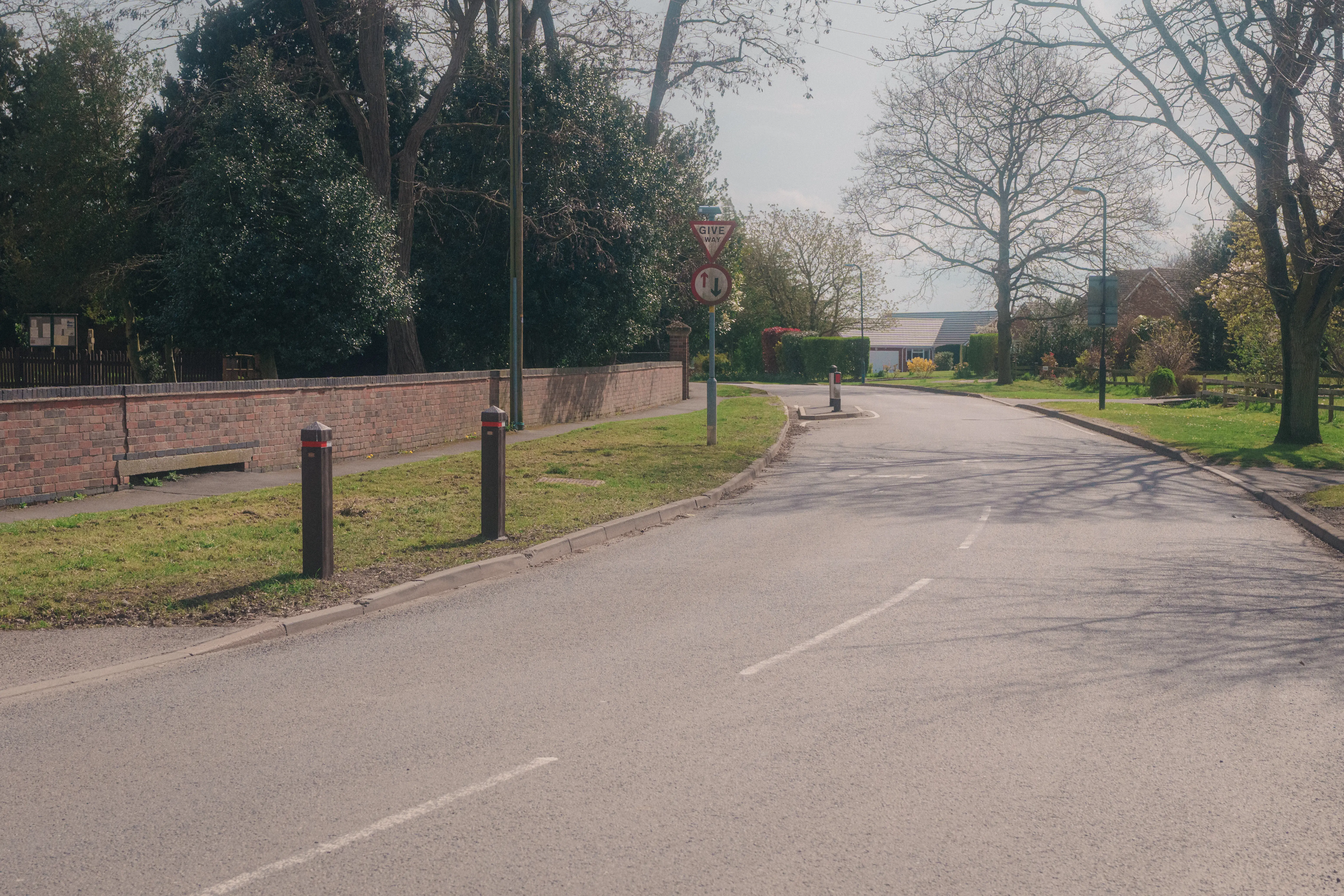 A winding road is lined with trees and bordered by a brick wall and grass on a sunny day.