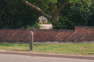A cracked brick wall stands in front of a tree-lined area, with a post and grassy verge along the roadside.