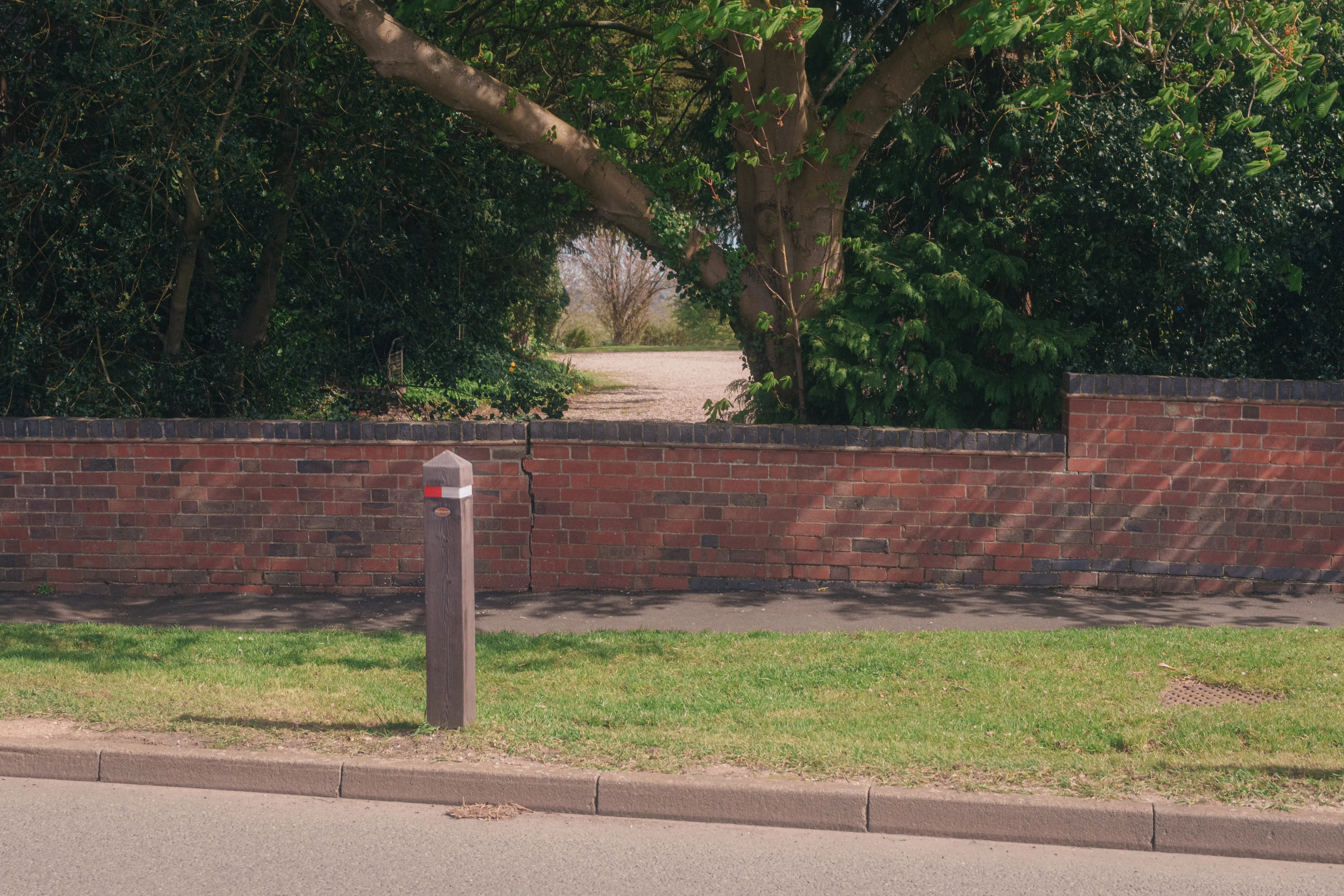 A brick wall with an opening surrounded by trees and a bollard in the foreground lines a sidewalk adjacent to a grassy area and a road.