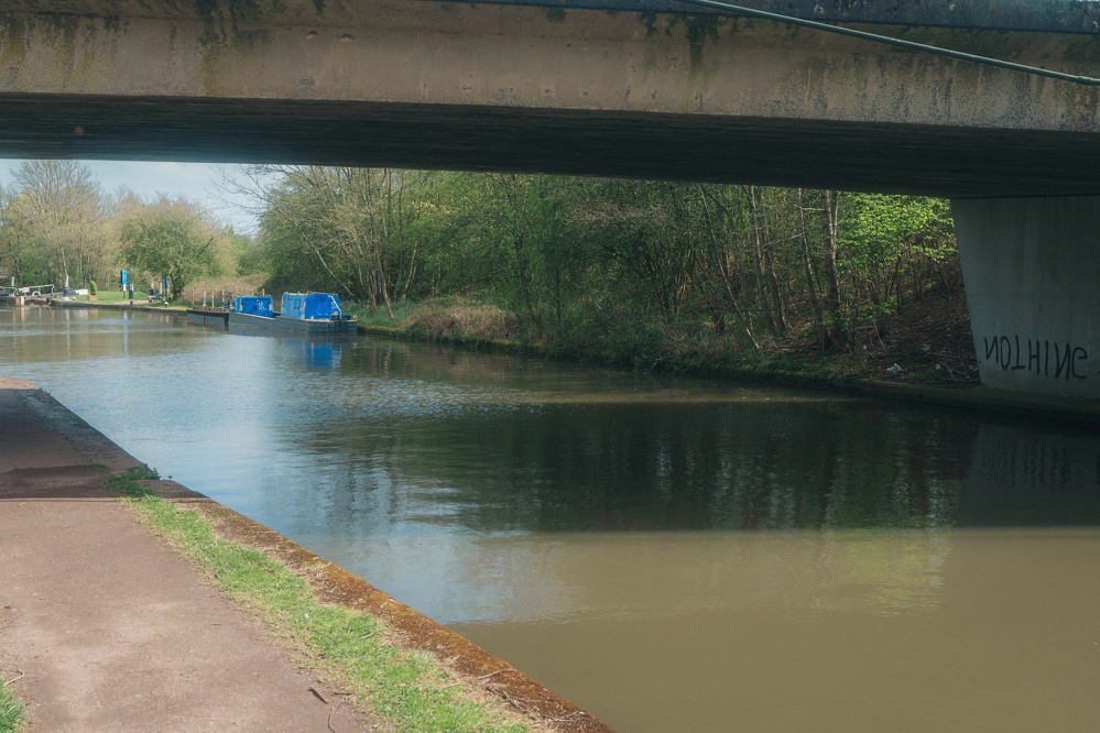 A canal with boats is partially shaded by a bridge, surrounded by greenery.