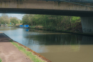 A canal with boats is partially shaded by a bridge, surrounded by greenery.