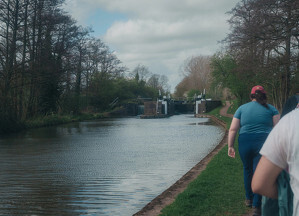 Two people are walking along a path beside a tranquil canal with a series of locks and surrounded by trees.