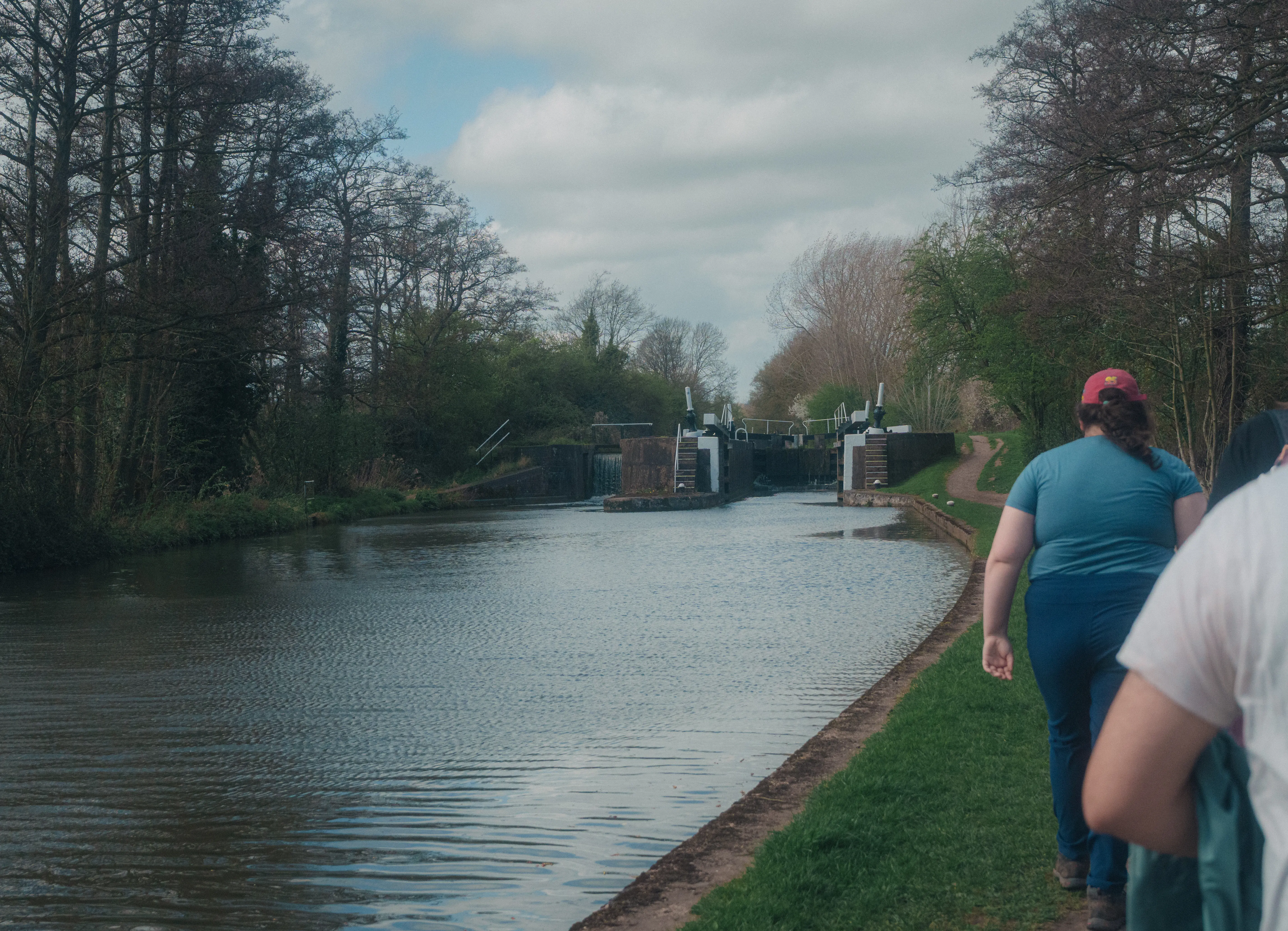 People are walking along a canal path lined with trees and a lock system in the distance.