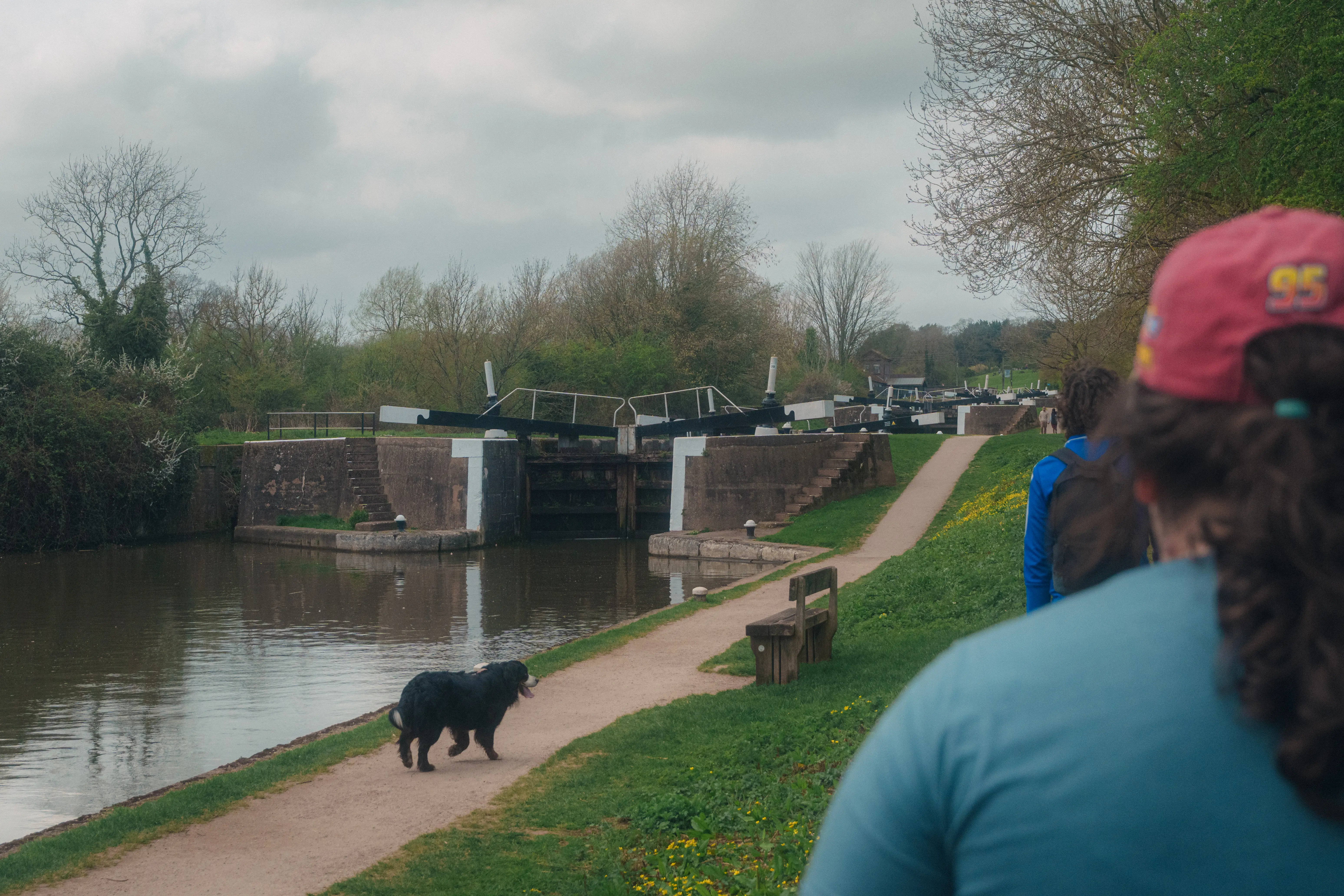 A person and a dog walk along a canal path next to a lock system on a cloudy day.