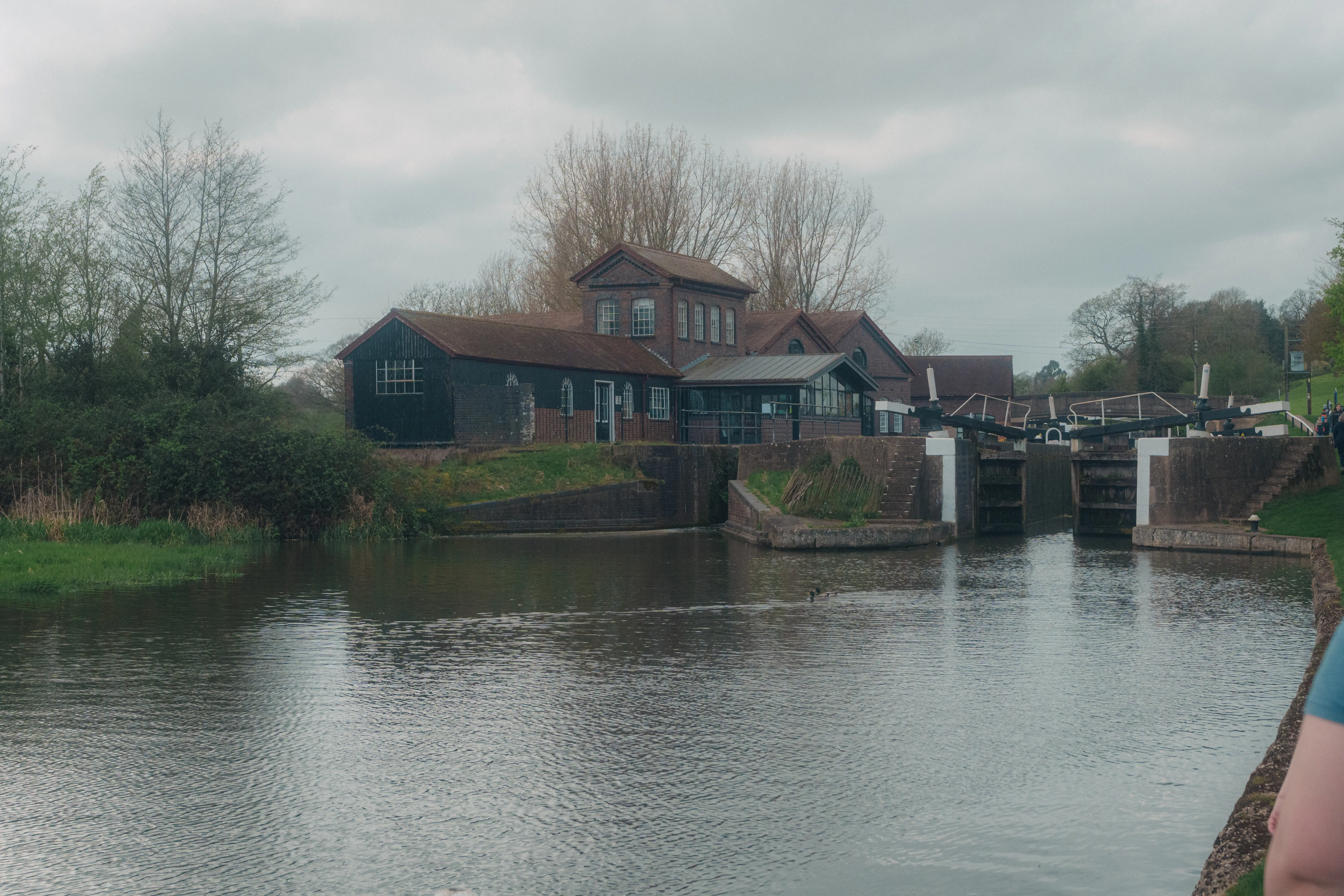 A quiet canal scene features a lock with adjacent buildings surrounded by trees under a cloudy sky.