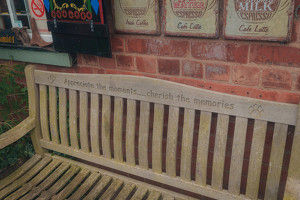 A wooden bench with the engraved message Appreciate the moments... cherish the memories is set against a brick wall adorned with vintage-style coffee signs.