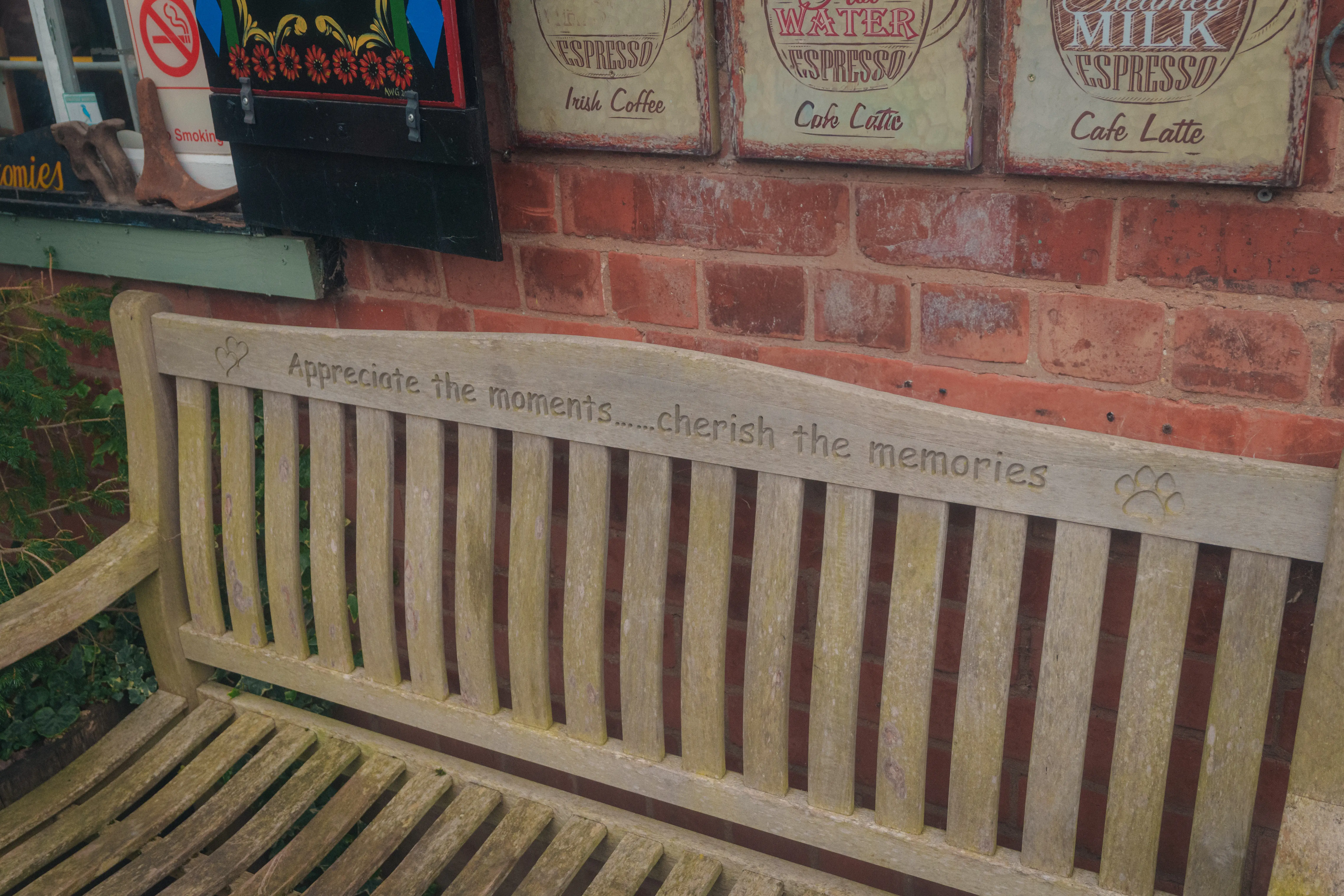 A wooden bench with the engraved message Appreciate the moments...cherish the memories is set against a brick wall decorated with vintage coffee signs.