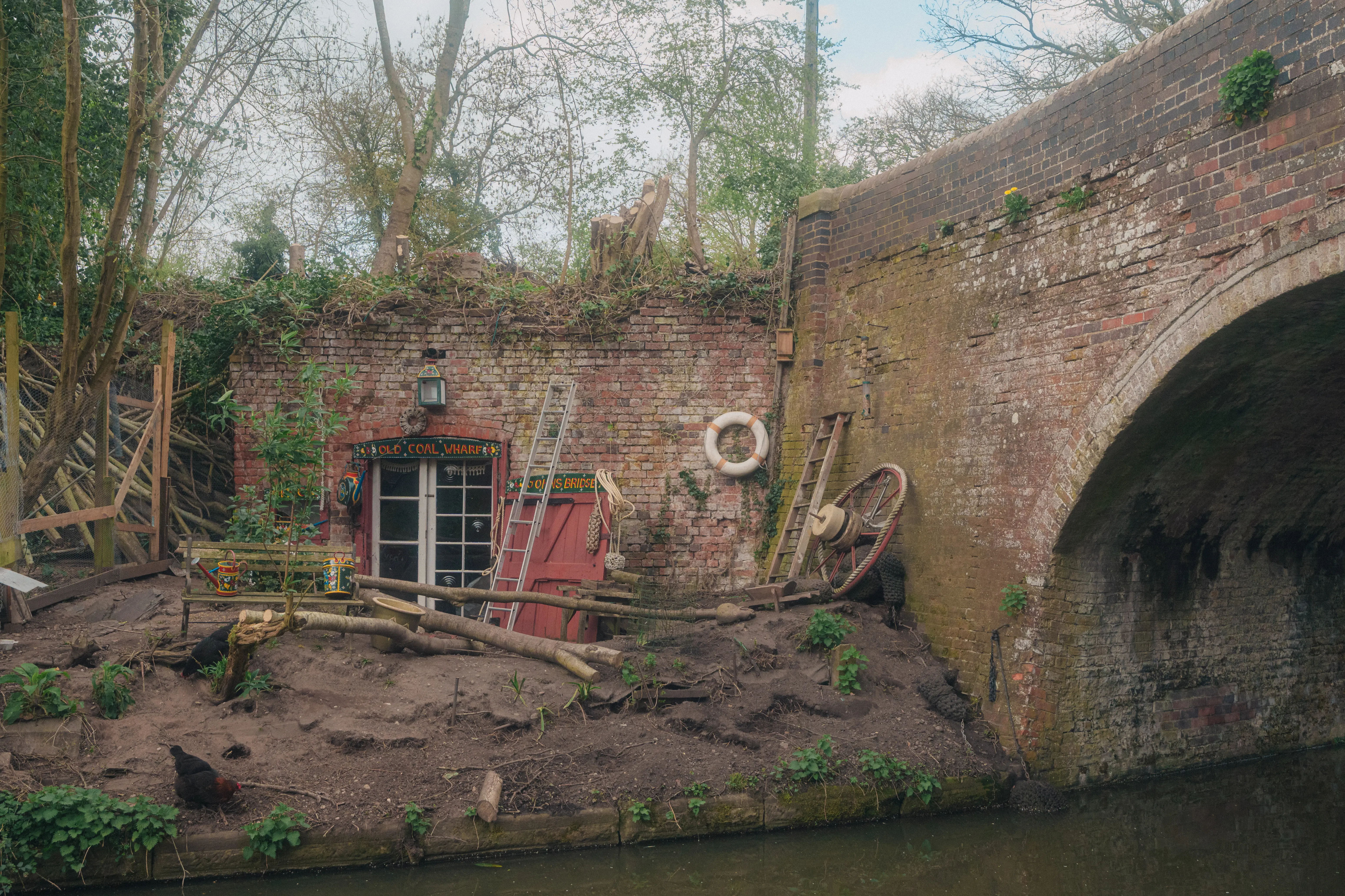 A quirky, Hobbit-like house is built into a canal bank next to a brick bridge, surrounded by various nautical and rustic decorations.