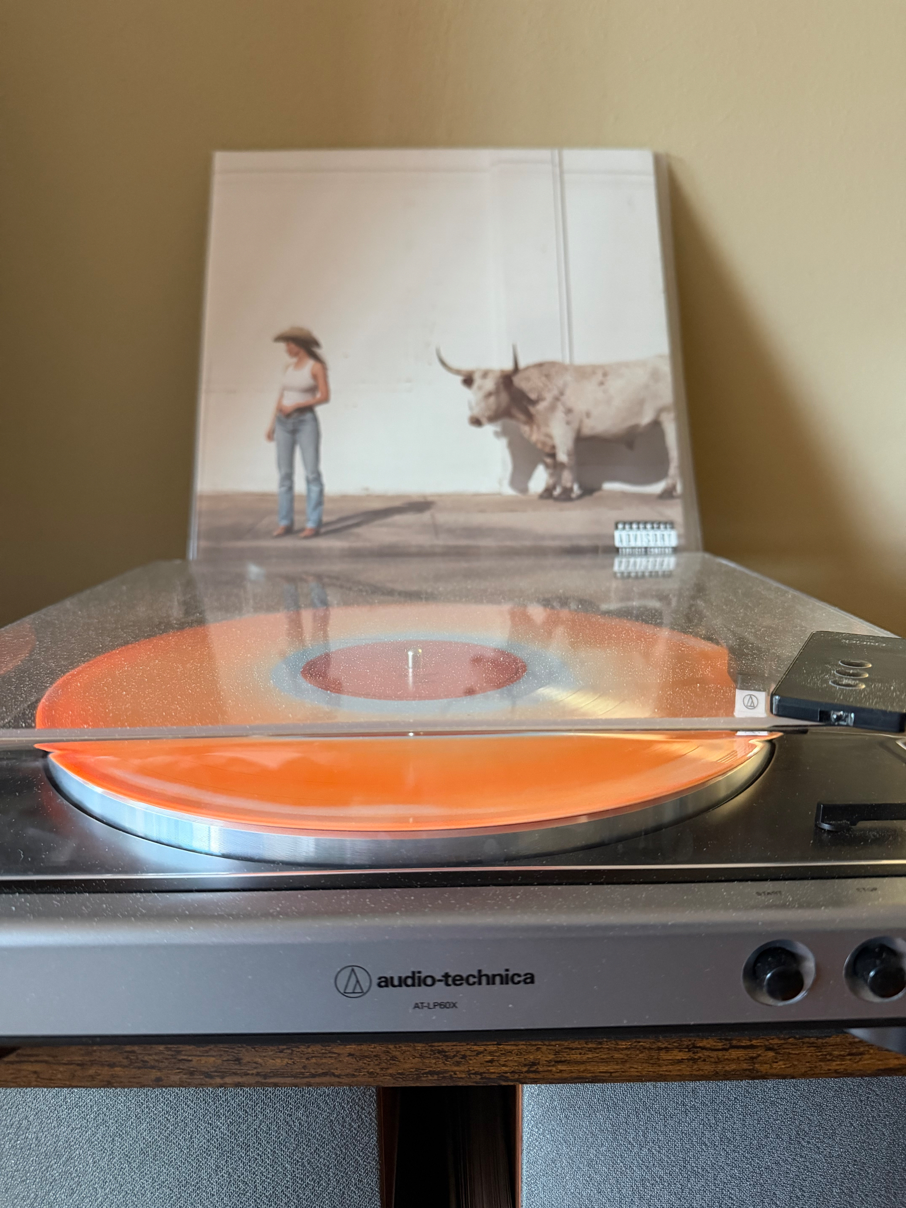 An orange vinyl record is spinning on an Audio-Technica turntable with an album cover featuring a person and a white longhorn in the background.