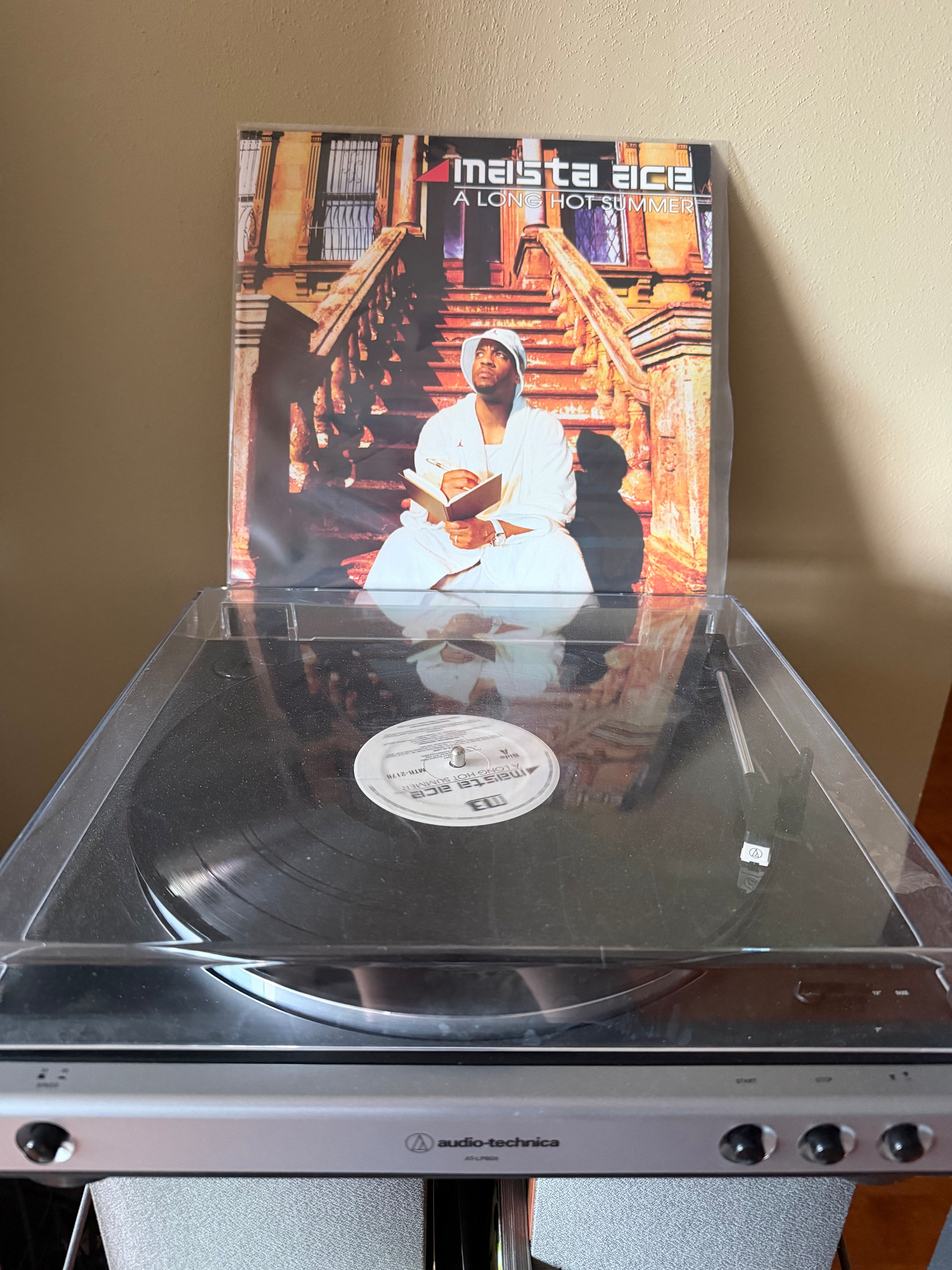 A vinyl record titled A Long Hot Summer by Masta Ace is on a turntable, with its album cover depicting a person sitting on steps holding a book.