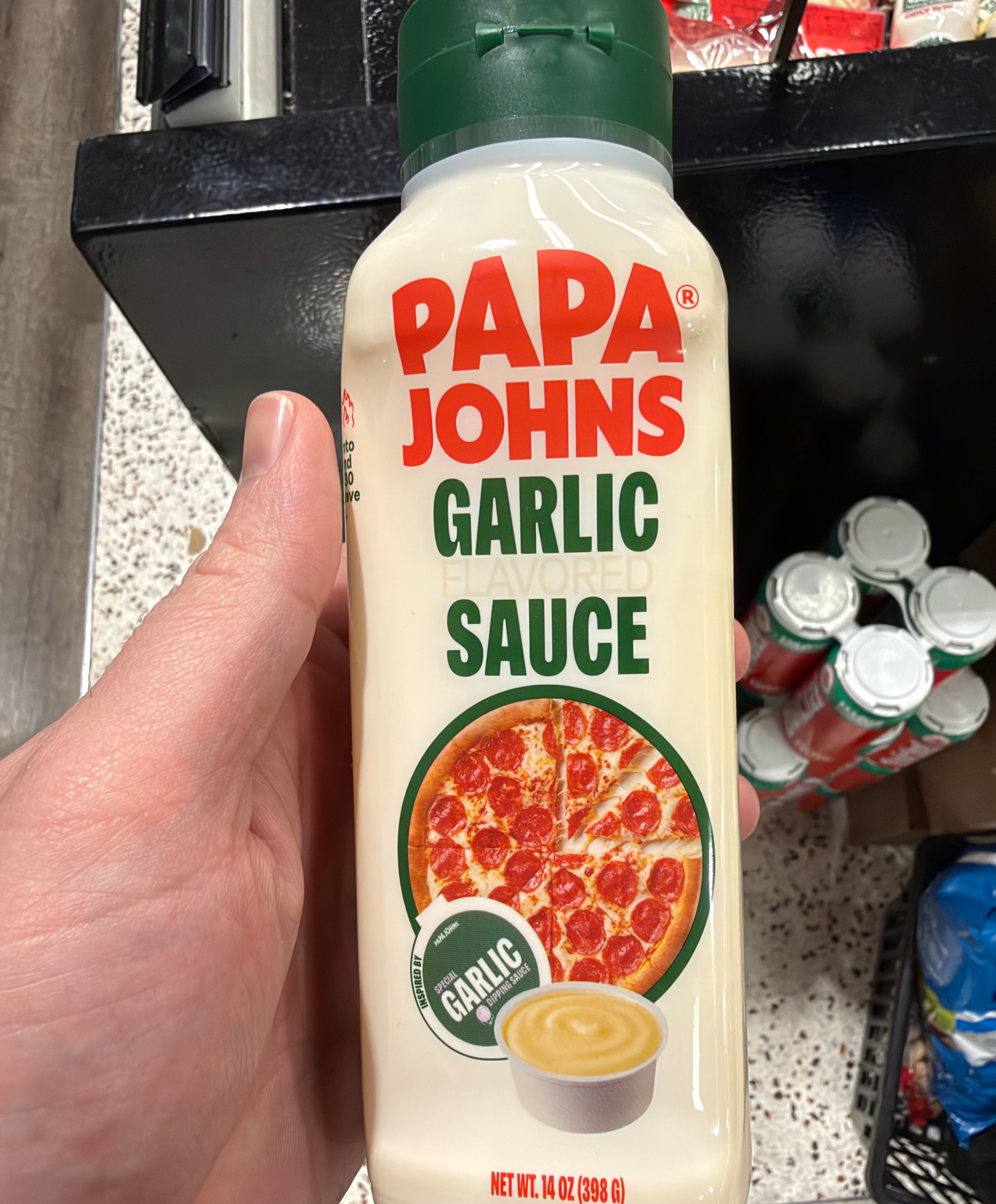 A bottle of Papa John's Garlic Sauce is being held in a hand, with the label featuring a pepperoni pizza and a small container of garlic sauce.