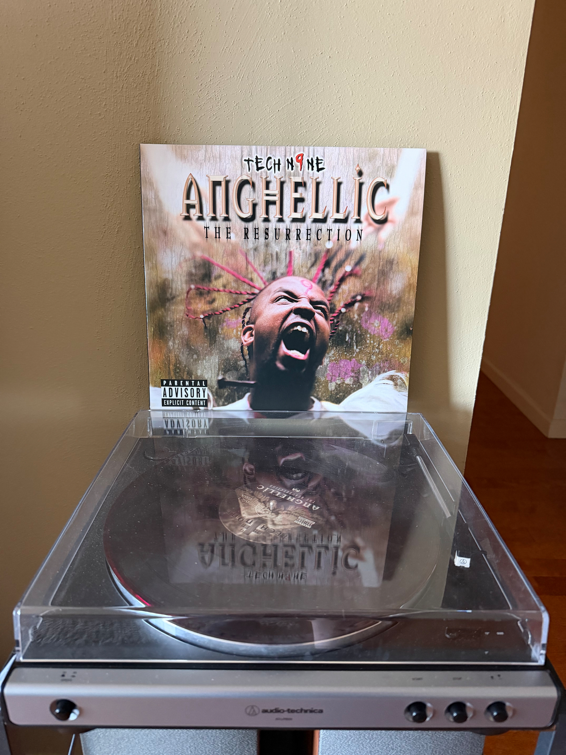 A vinyl record player with a clear dust cover displays the album cover of Tech N9ne's Anghellic: The Resurrection, showing a close-up of the artist with braided hair and an expressive face.
