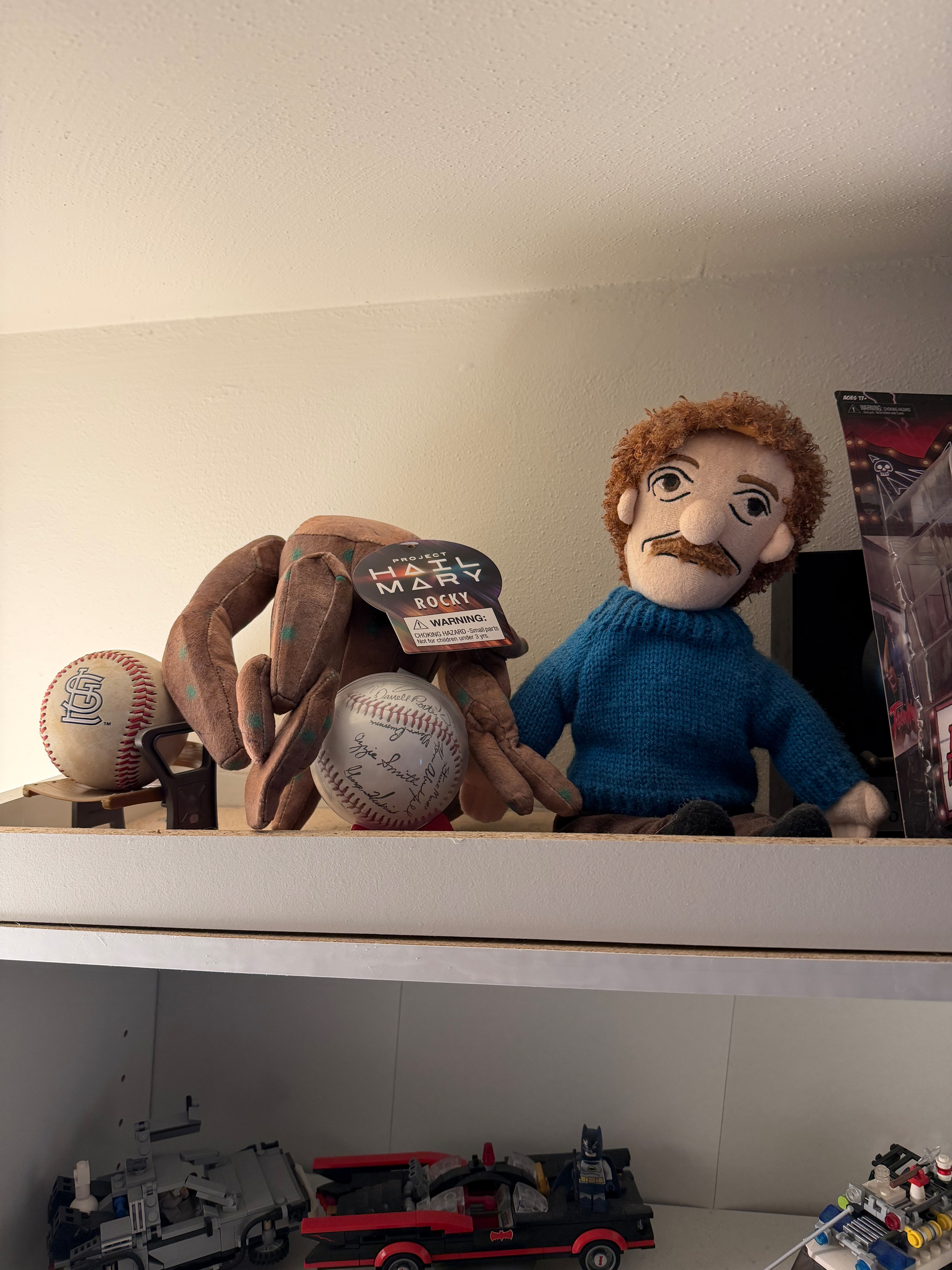 A shelf displays a plush doll of Rocky from Project Hail Mary next to a plush doll or Kurt Vonnegut, a baseball glove holding baseballs, and some toys, including a vehicle model.