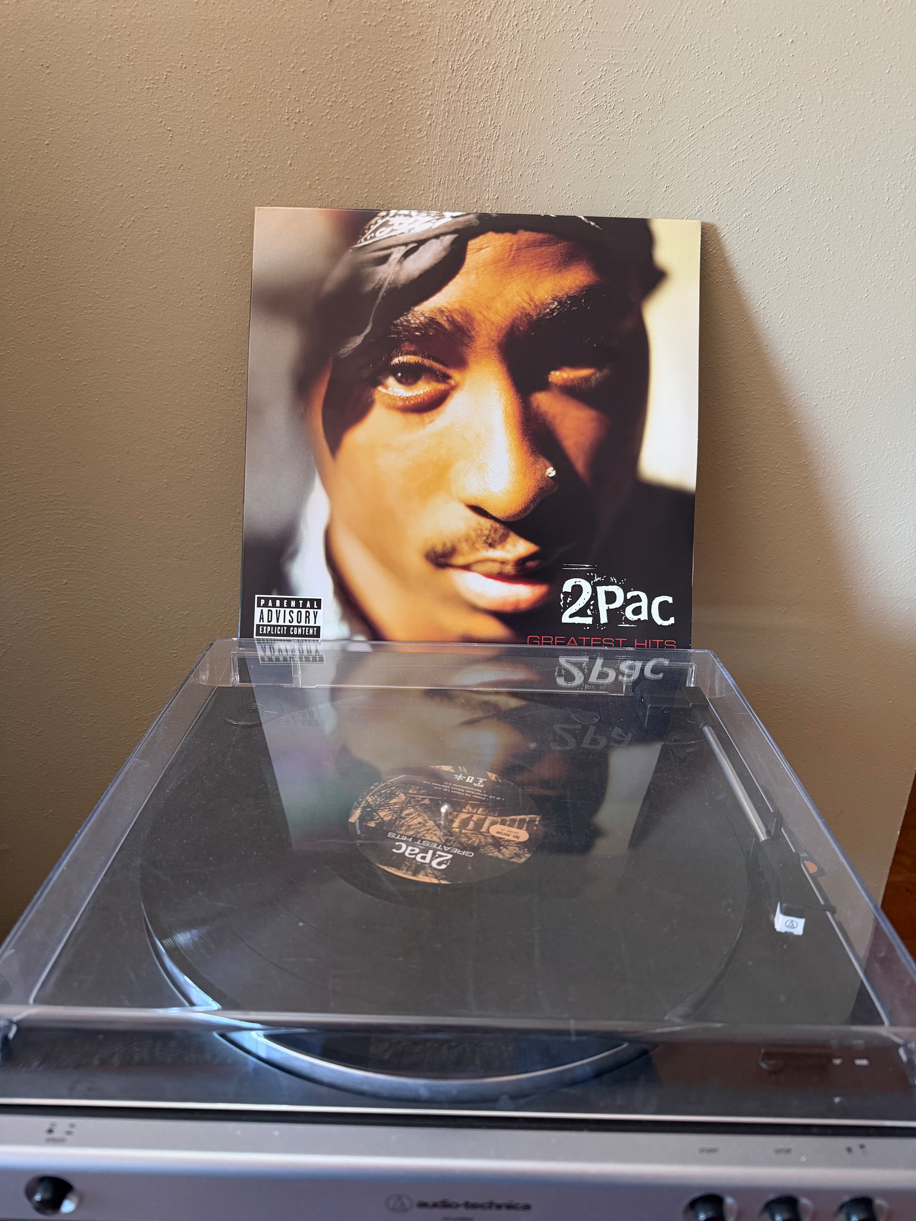 A vinyl record with the cover of a music album, featuring a close-up portrait of a man, is placed on a record player. 2pac greatest hits on vinyl. 