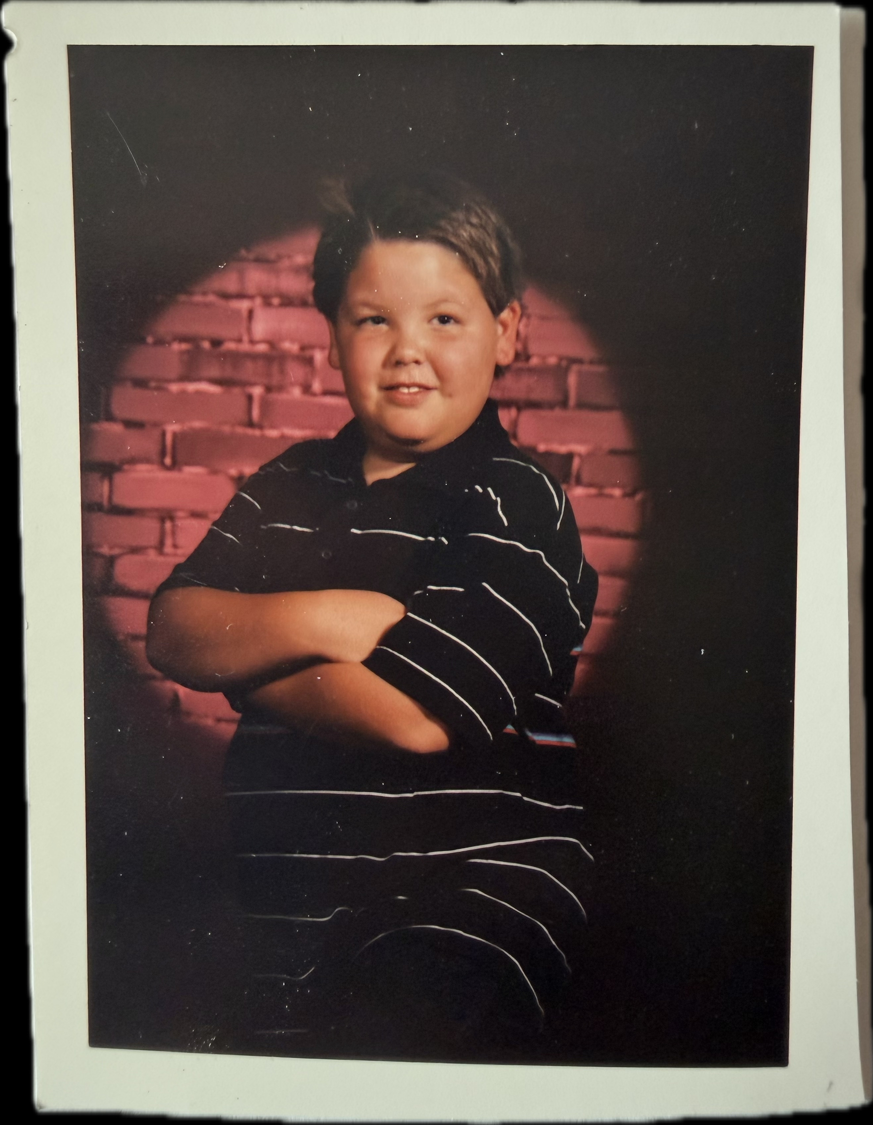 A young boy with crossed arms smiles in front of a red brick background.