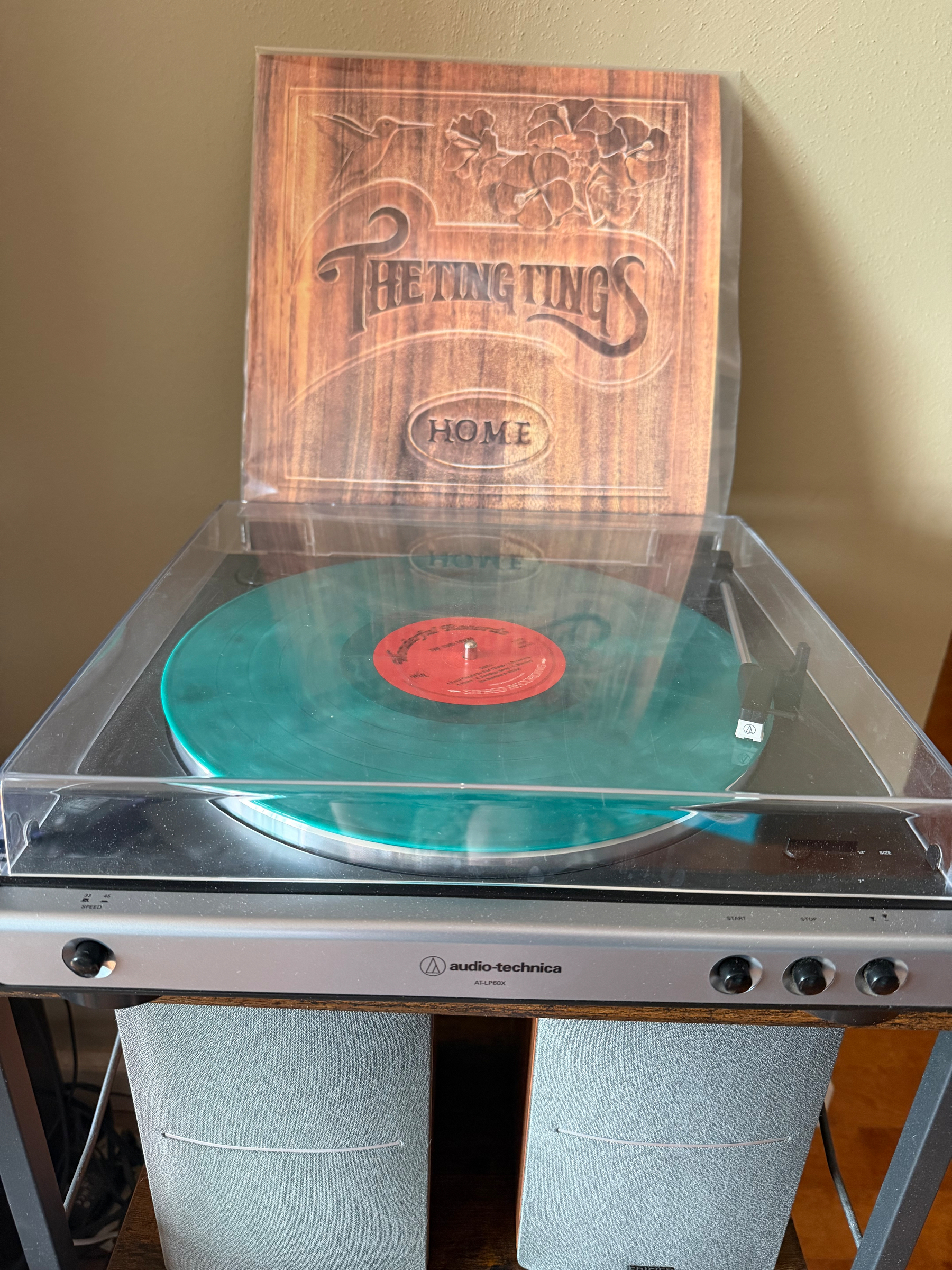 A vinyl record is playing on an Audio-Technica turntable with an album cover displayed behind it. The album is Home by the Ting Tings. 