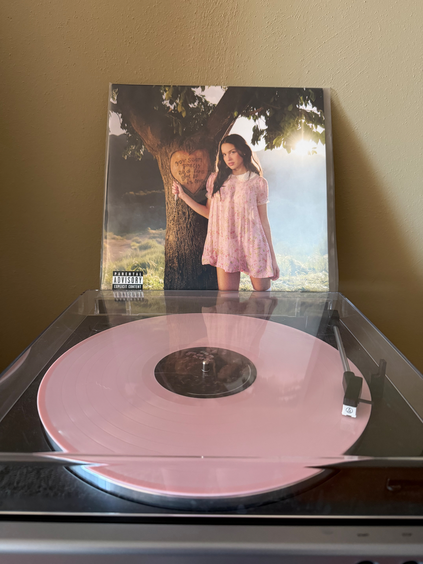 A pink vinyl record is spinning on a turntable with an album cover featuring a woman in a dress standing by a tree in the background. Olivia Rodrigo- you seem pretty sad for a girl so in love