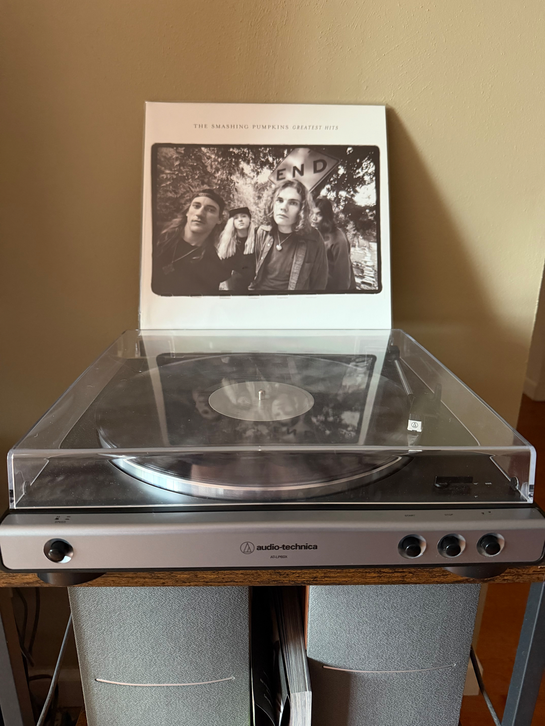 A vinyl record player with a clear dust cover is set up on a wooden stand displaying The Smashing Pumpkins Greatest Hits album cover.