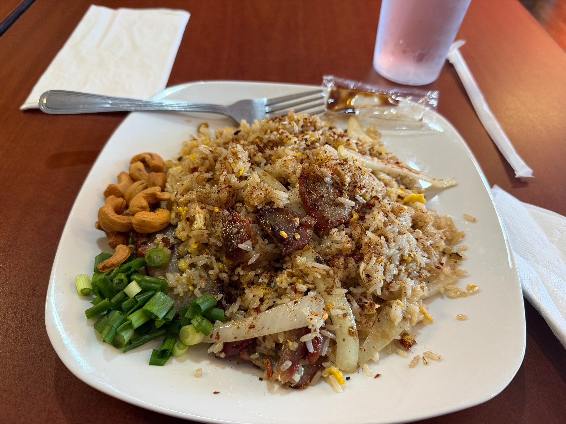 A plate of fried rice garnished with green onions, cashews, and served with a fork, is on a wooden table alongside a glass and a napkin.