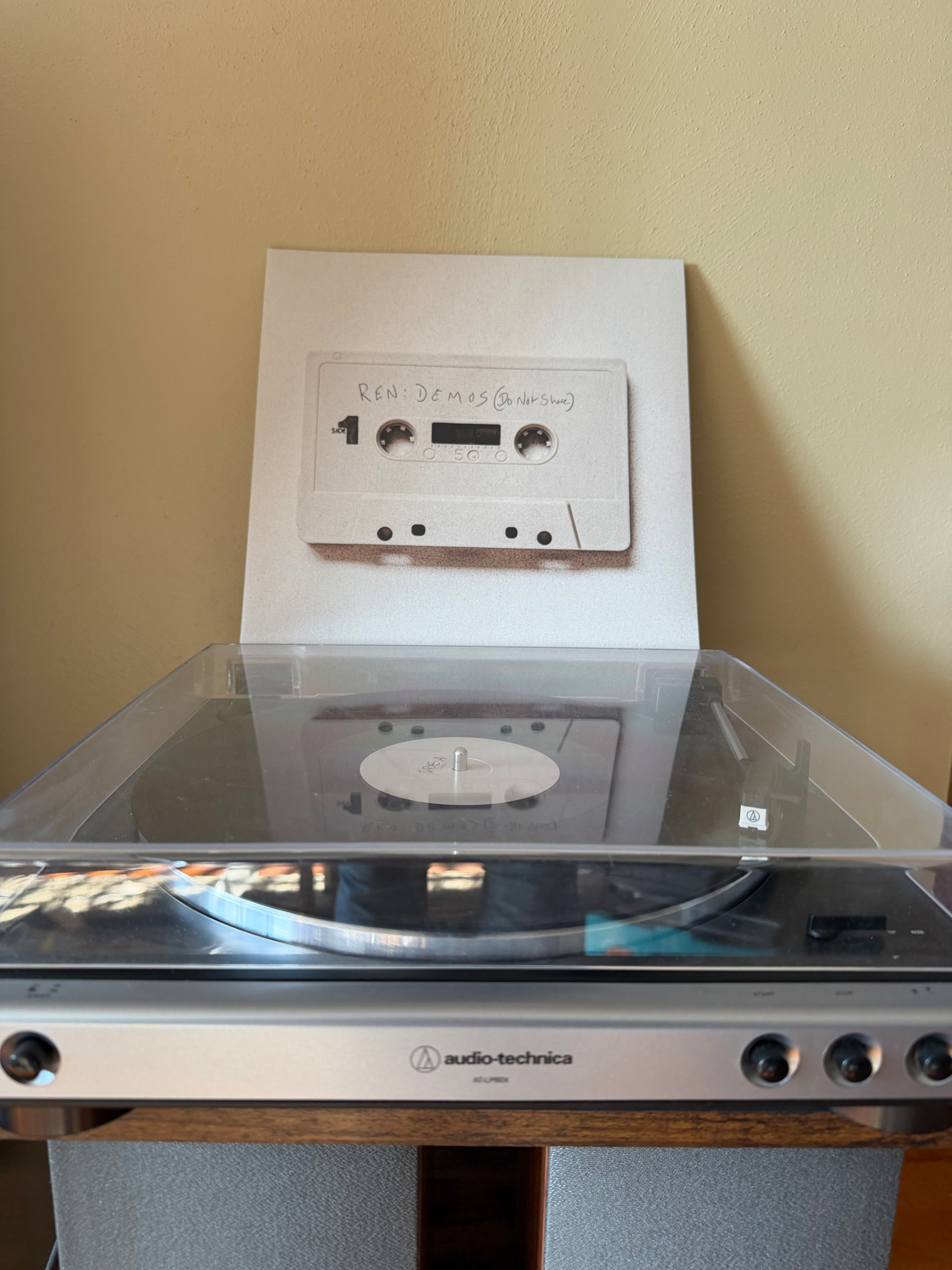 A vinyl record player with a clear cover is positioned in front of an artwork depicting a cassette tape labeled REM DEMOS (unreleased).”
