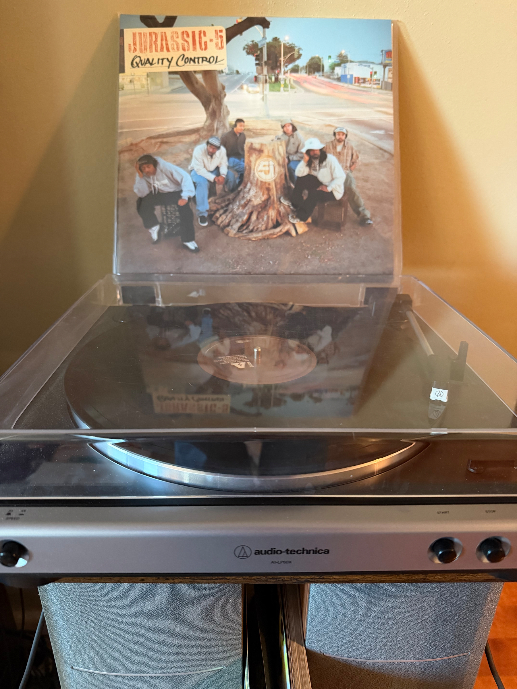 A vinyl record player is spinning with an album cover of a group titled Jurassic 5 Quality Control displayed behind it.