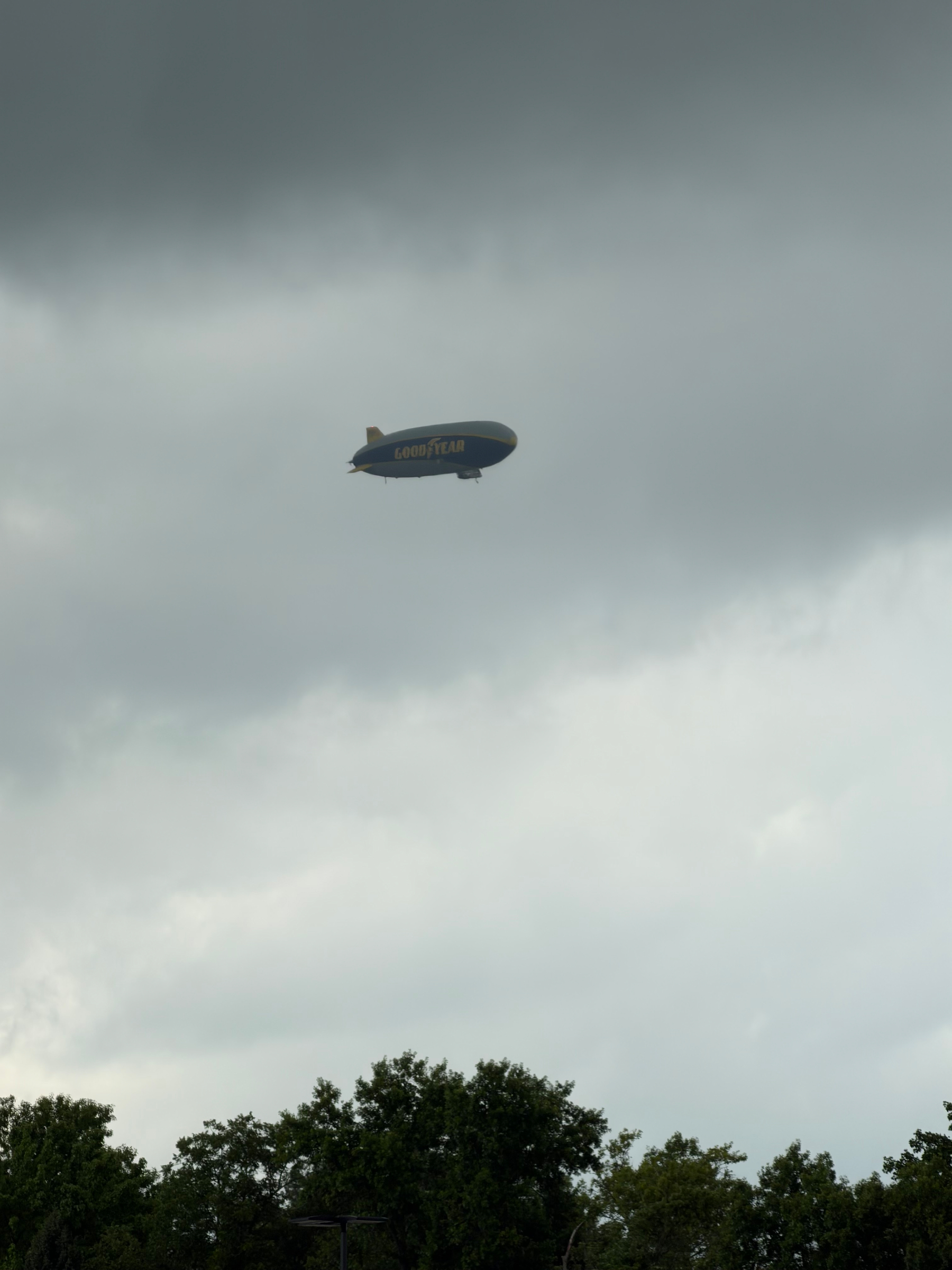 A Goodyear blimp is flying high in a cloudy sky above a treeline.