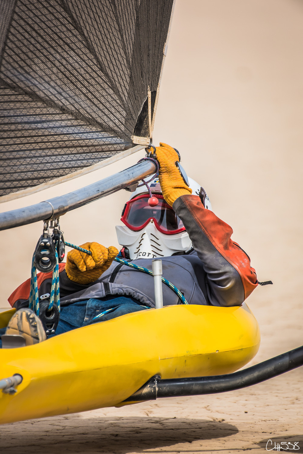 A person is riding a sand yacht on a sandy surface, wearing protective gear and gloves while gripping the sail.