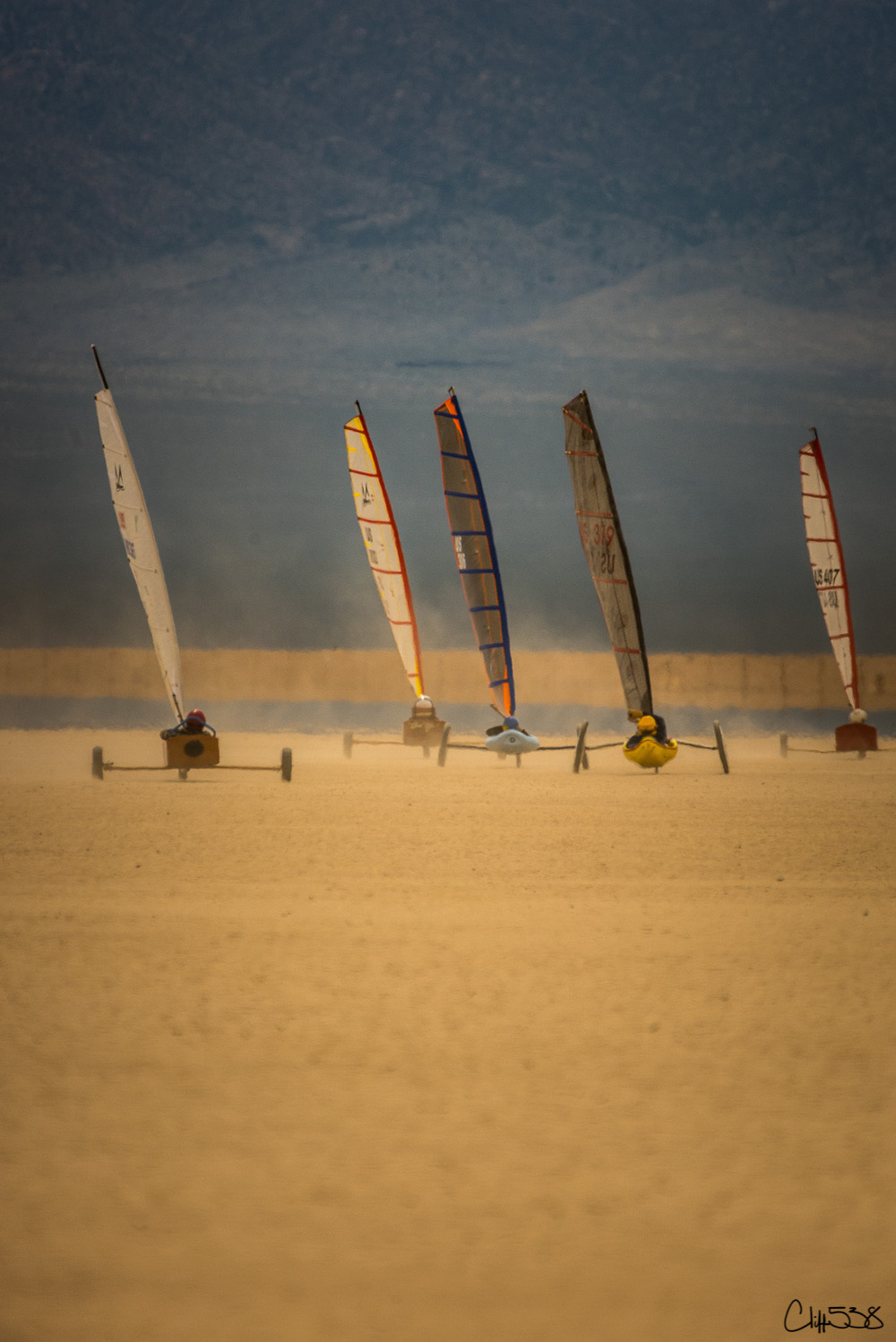 Five land yachts with colorful sails race across a dusty, desert landscape.