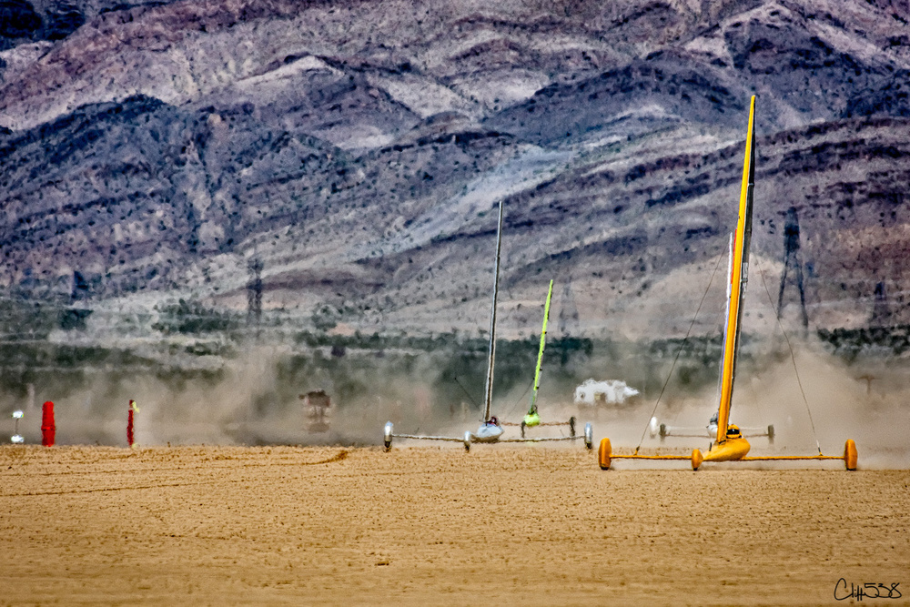 A series of brightly colored land yachts race across a dusty desert landscape with mountains in the background.