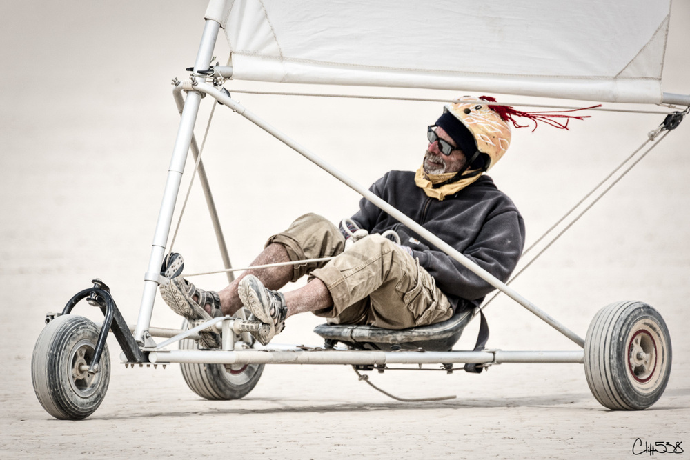 A person wearing a helmet and casual clothing is sitting in a small, open-frame land sailing vehicle on a flat surface.