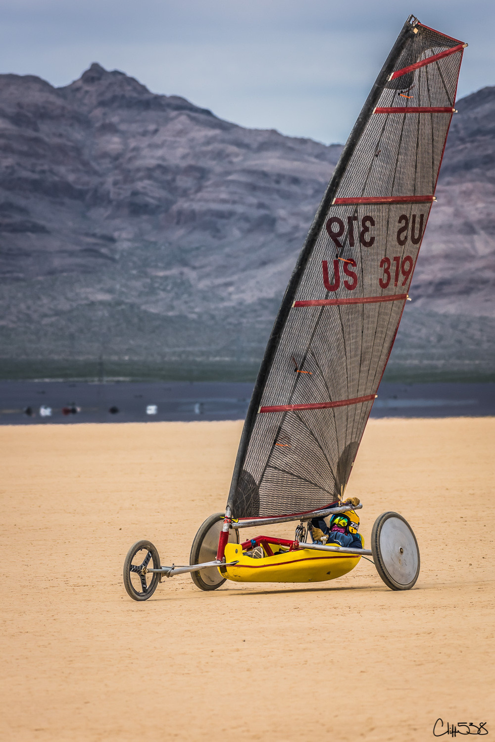 A land sailor with a large sail navigates across a dry lake bed under a cloudy sky, with mountains in the background.