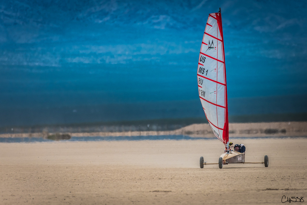 A land sailing vehicle with a large sail is speeding across a vast, sandy terrain under a clear blue sky.