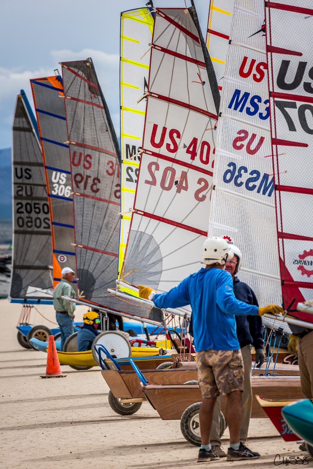 Several land sailing vehicles with colorful sails are lined up along a sandy shoreline, piloted by people in safety gear.