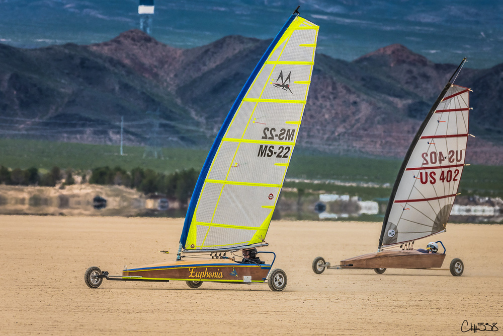 Two land sailboats are racing across a dry landscape with mountains in the background.