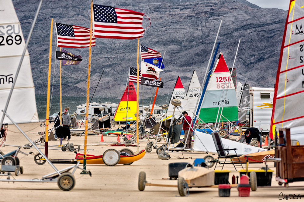 A group of colorful land yachts with American flags are gathered on a dry lake bed, surrounded by an arid landscape and RVs in the background.