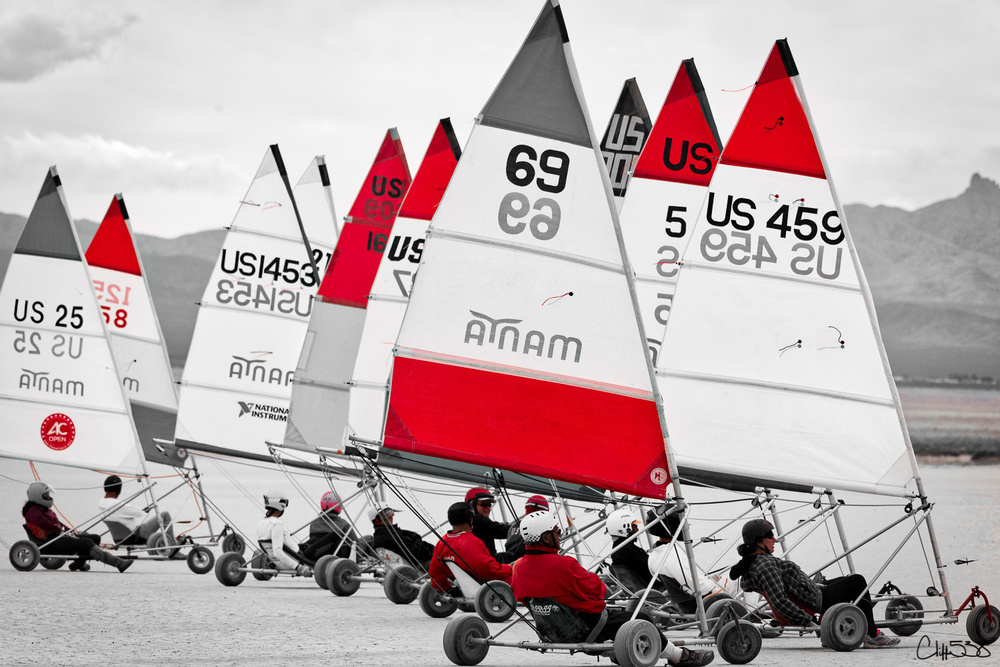 Several people are racing land sailboats with colorful sails across a flat, open terrain.