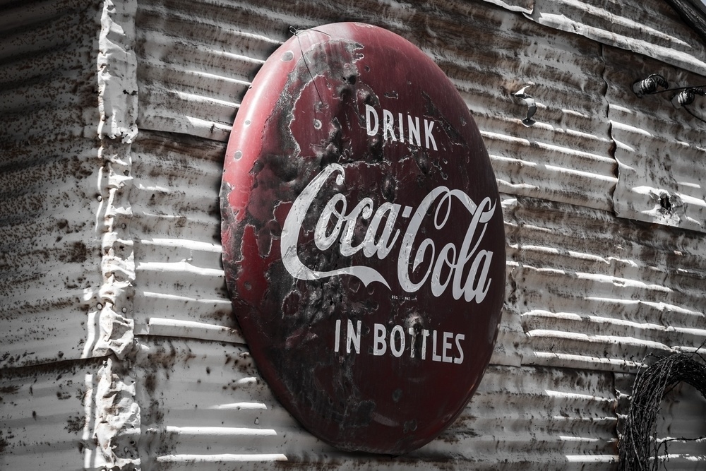 A weathered, vintage Coca-Cola sign displaying Drink Coca-Cola in Bottles is mounted on a rustic, corrugated metal wall.