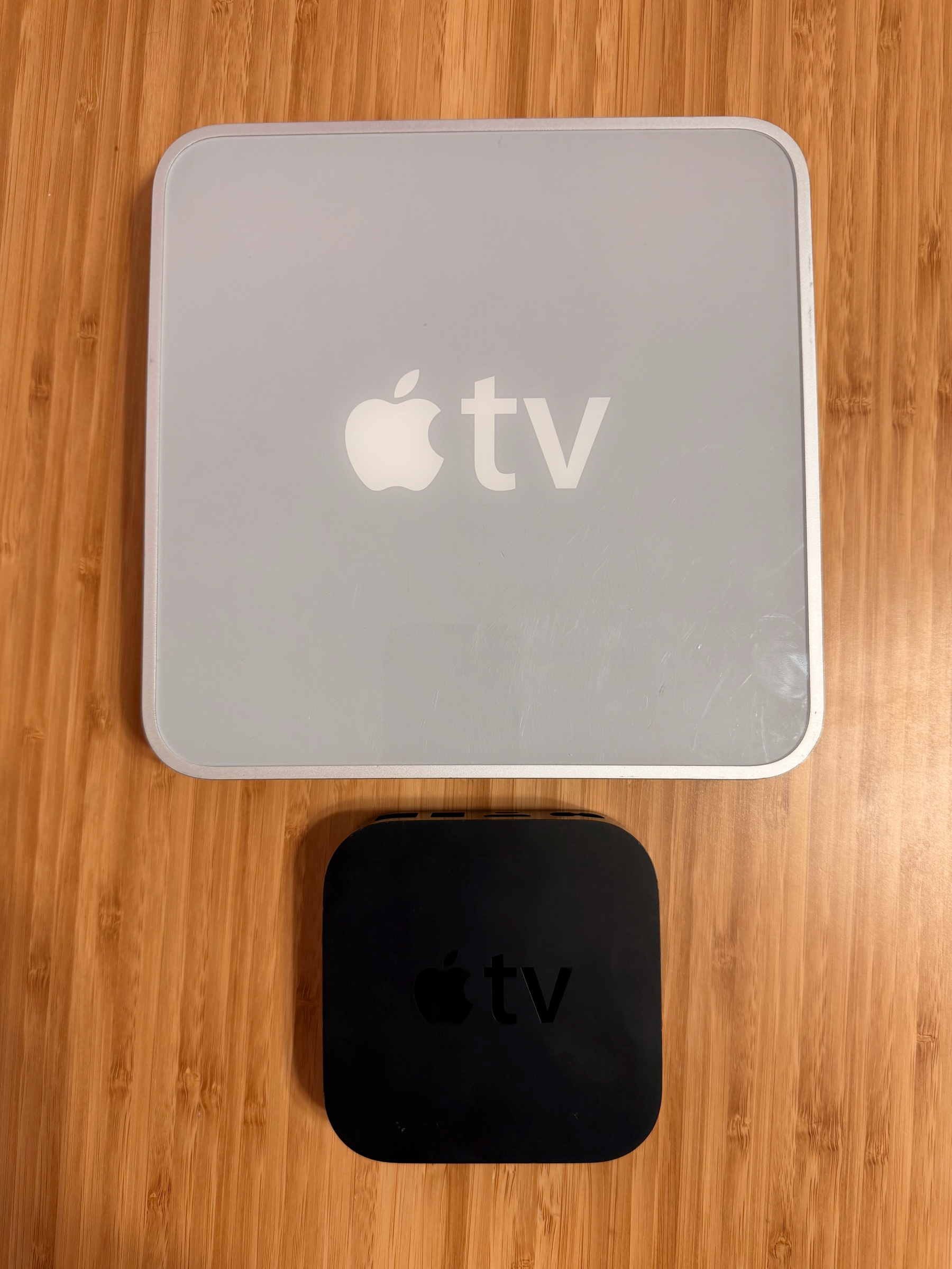 A small black Apple TV device is placed below a larger white Apple TV package on a wooden surface.