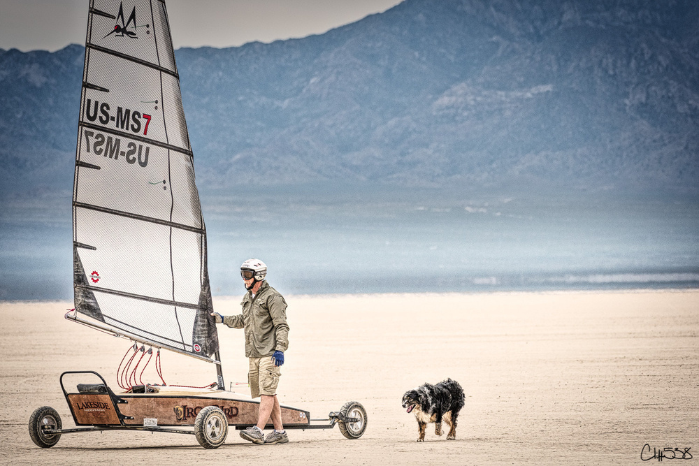 A person with a helmet and a dog stand beside a small land sailboat on a vast, dry landscape with mountains in the background.