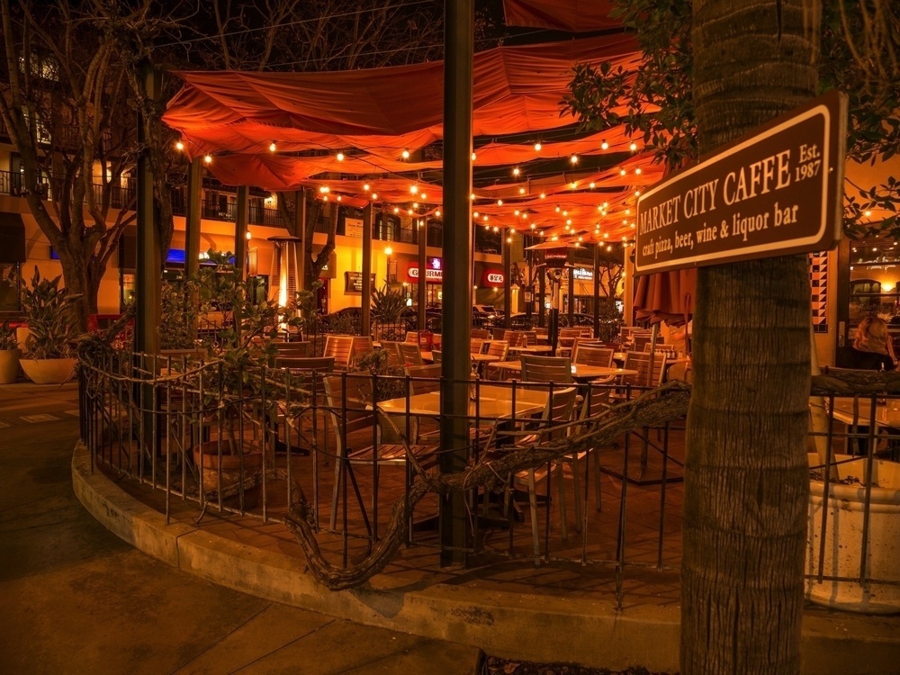 A warmly lit outdoor café patio is decorated with string lights and surrounded by empty tables and chairs.