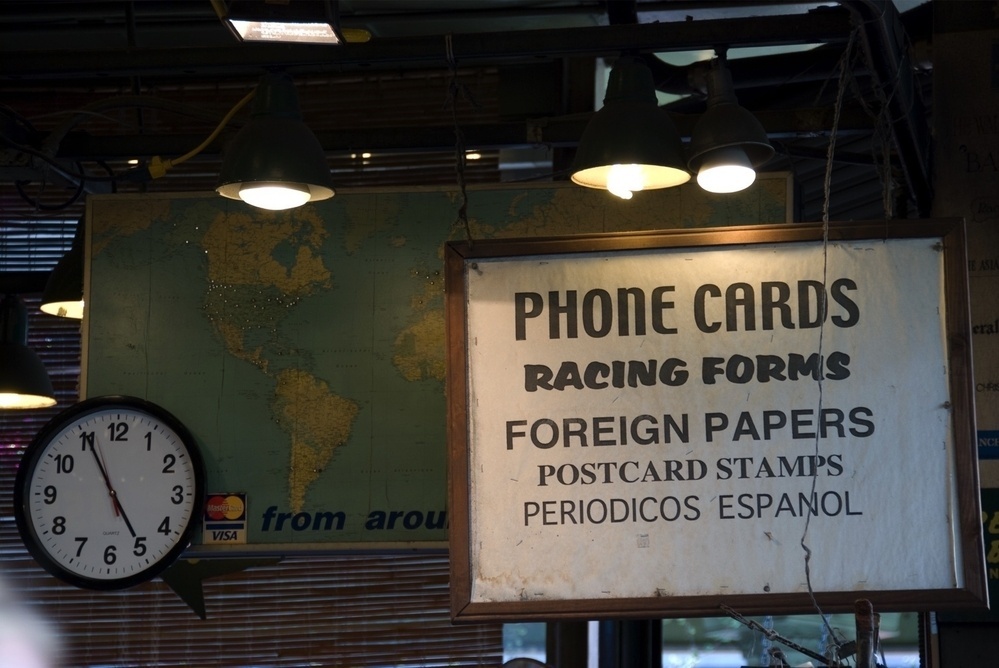 A store display features a variety of signs offering phone cards, racing forms, and foreign papers alongside a world map and a clock.