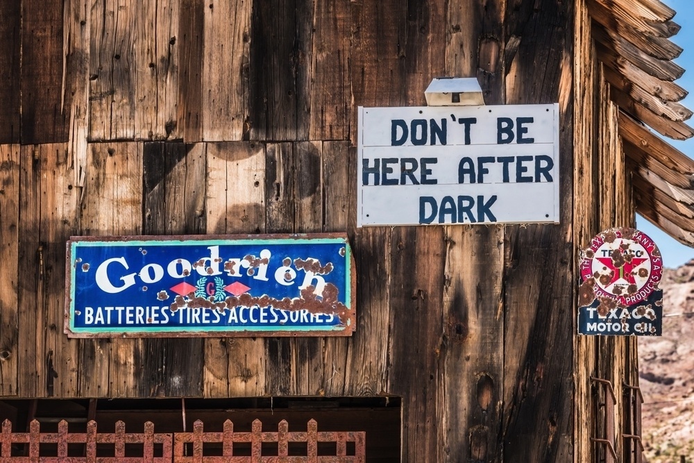 A weathered wooden building features vintage signs, including one that warns DON'T BE HERE AFTER DARK and advertisements for Goodrich and Texaco motor oil.