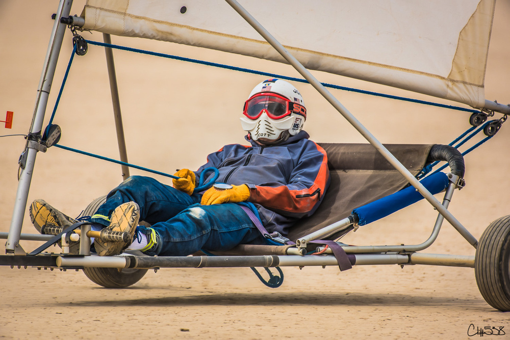 A person is sitting in a land sailing buggy wearing protective gear, including a helmet and goggles, on a sandy surface.